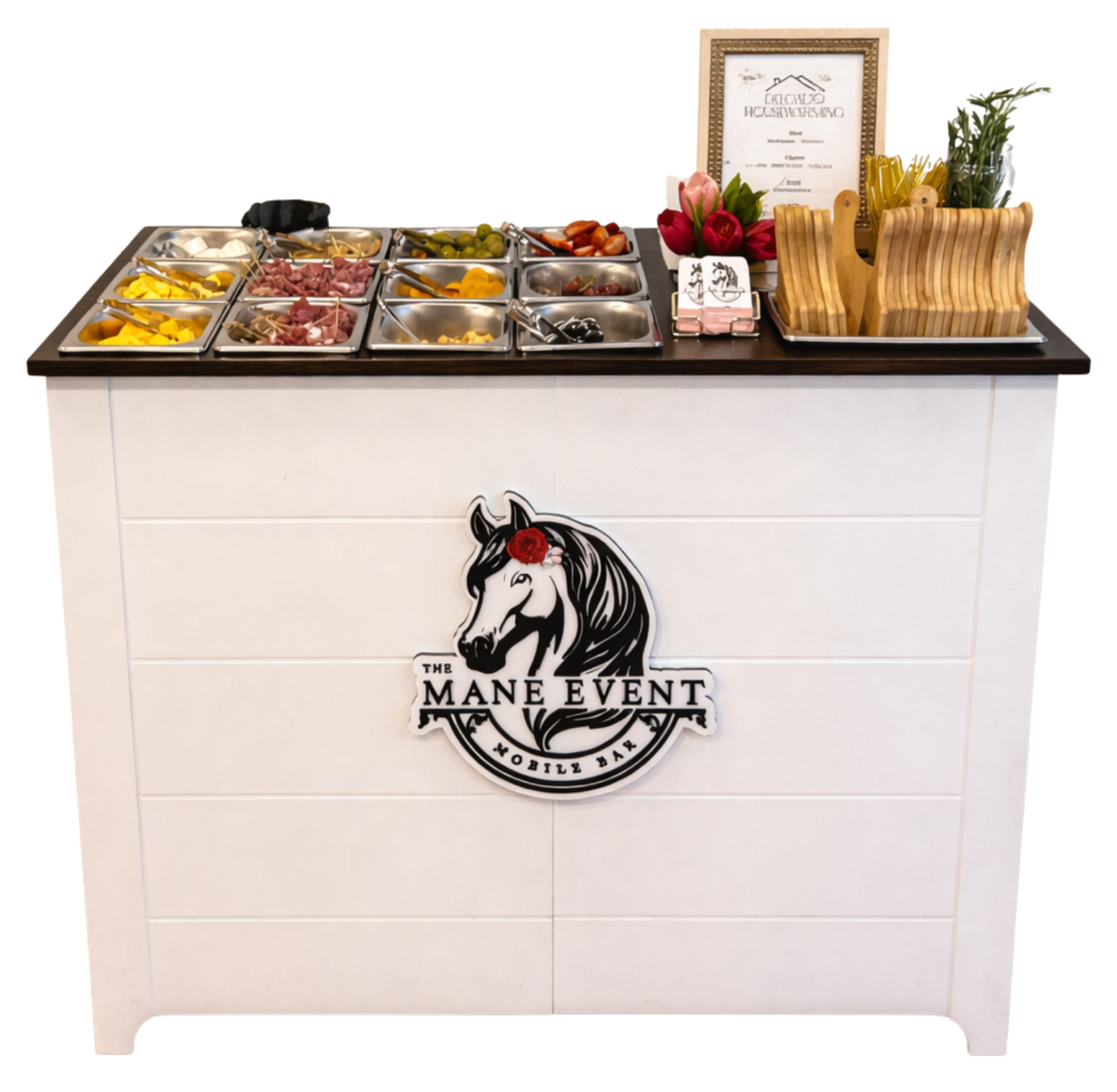 A white mobile bar with a black countertop displaying a variety of condiments and toppings in metal containers, a framed certificate, a small stack of pink napkins, and a container with yellow utensils and some greenery. The front of the bar has a logo featuring a black and white horse head with a red flower and text that reads "The Mane Event Mobile Bar."