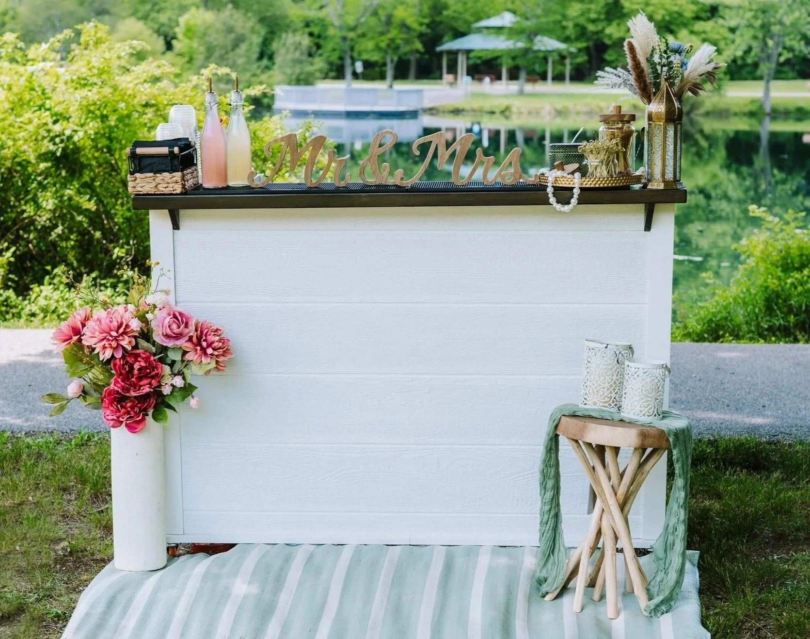 Decorative outdoor wedding bar setup with a white wooden backdrop, pink and green floral arrangement, and various drinks and accessories on top, with a lake and gazebo in the background.