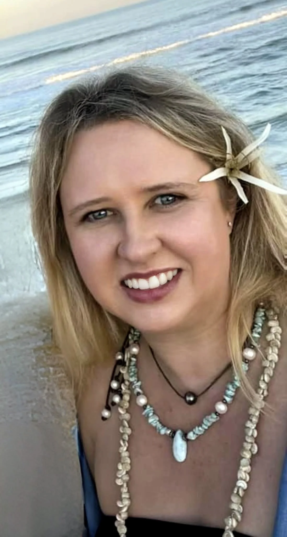A woman with blonde hair wearing a flower in her hair, leather and pearl necklace, and a blue top, smiling on the beach with the ocean behind her. This is Jennifer Russell, owner of Blue Palm Travel.