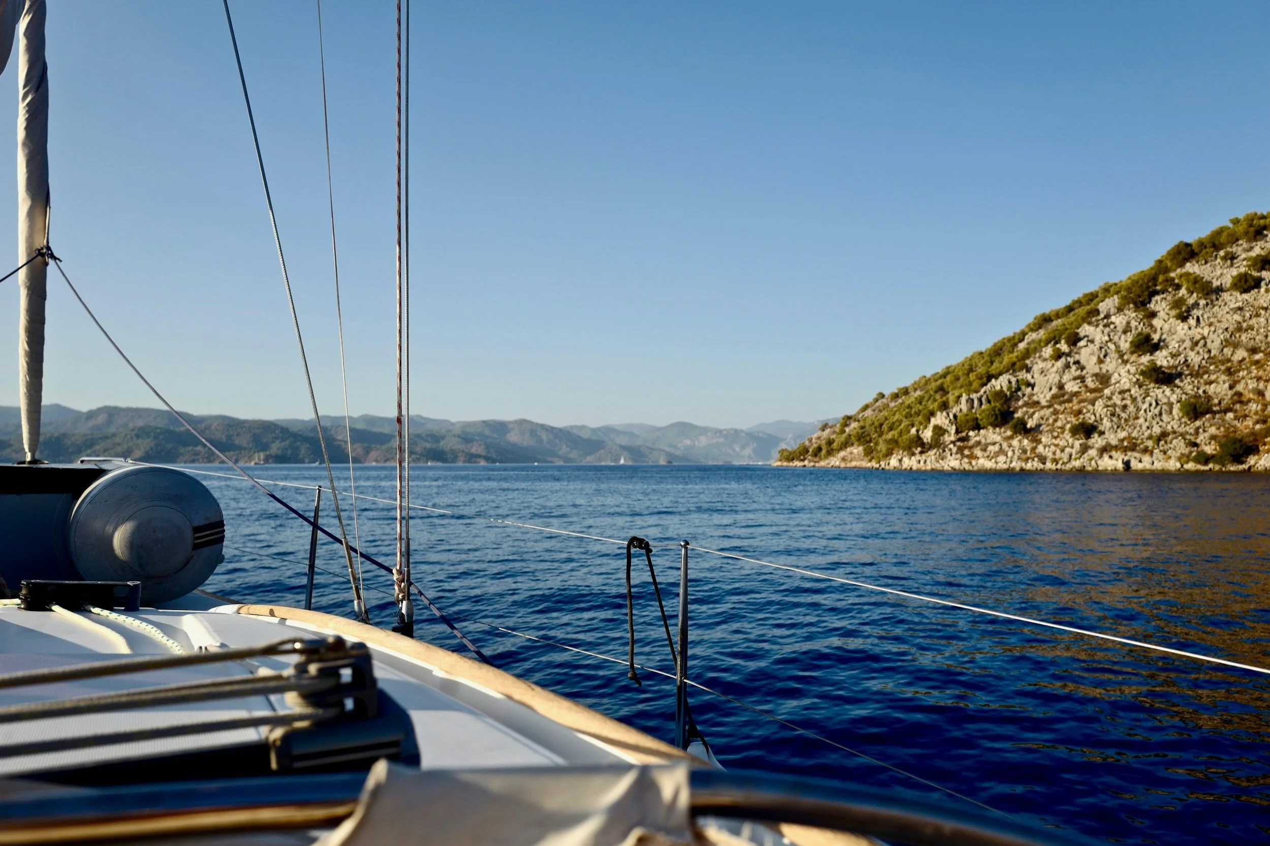 View of a calm blue sea from a sailboat with mountains on the horizon and a hillside on the right, clear sky overhead.