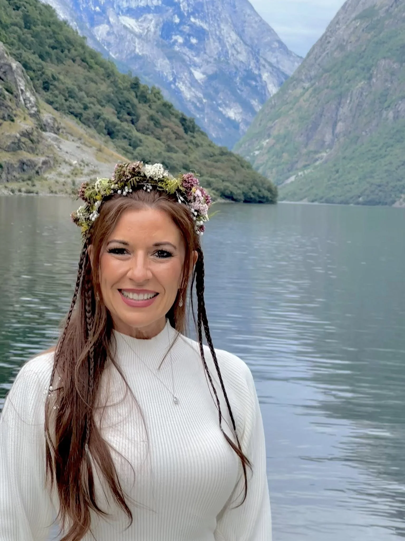 A woman with long brown hair, wearing a white sweater and a flower crown, smiling in front of a lake surrounded by green mountains.