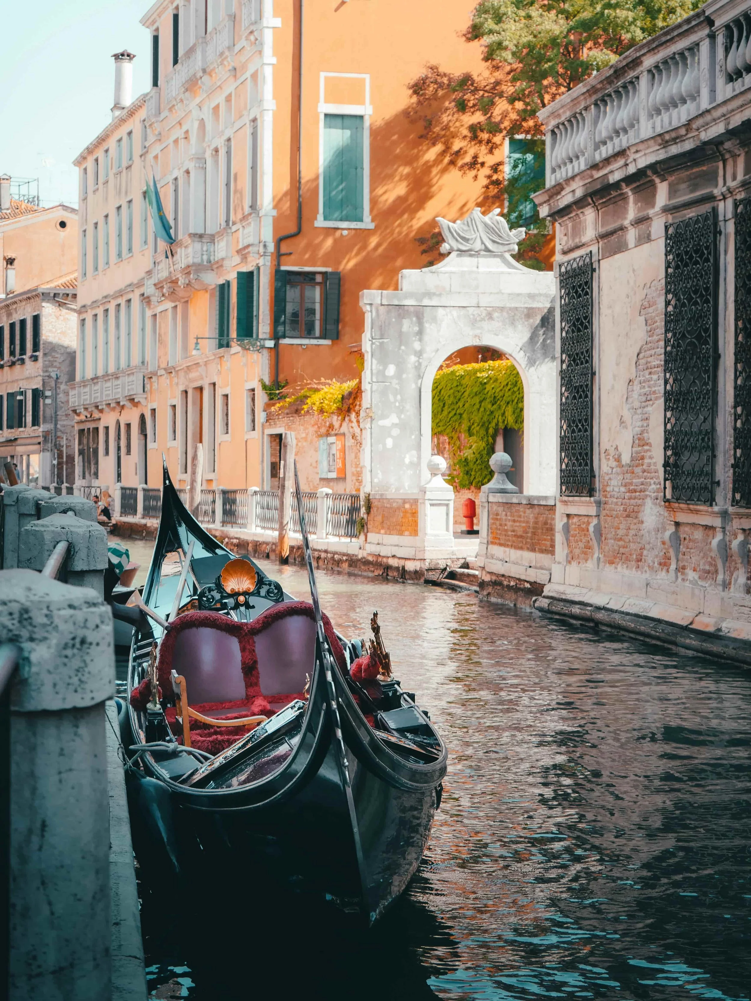 Gondola docked along a canal with colorful buildings, a white archway, and trees in Venice, Italy.  A Luxury, Custom Adventure you will love when your travel is created by Blue Palm Travel, Jennifer Russell