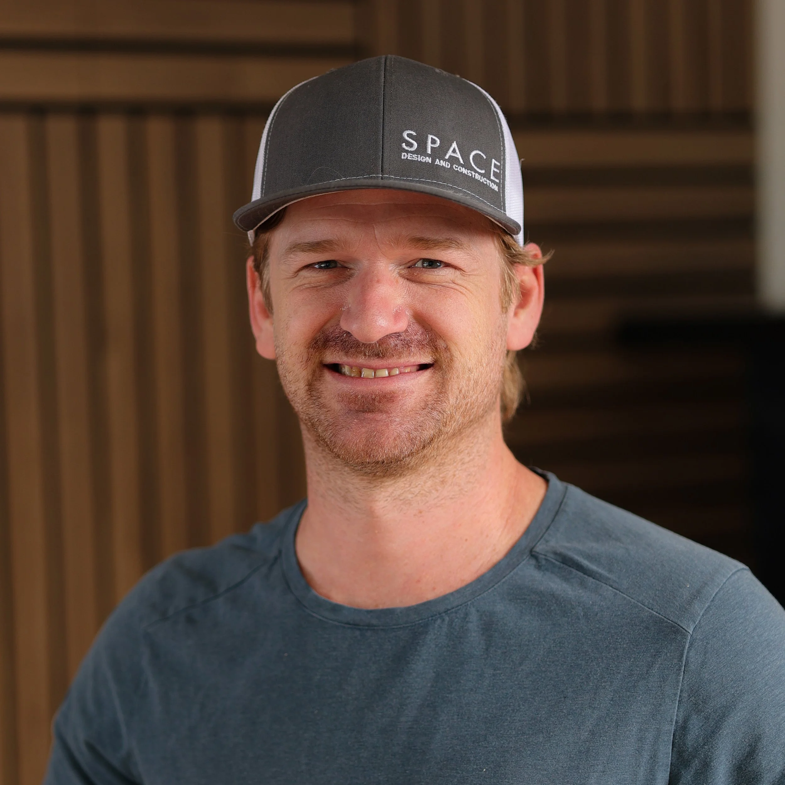 Close-up of a smiling man wearing a gray baseball cap with white embroidery that reads 'SPACE DESIGN AND CONSTRUCTION', and a gray t-shirt, with a wooden background.