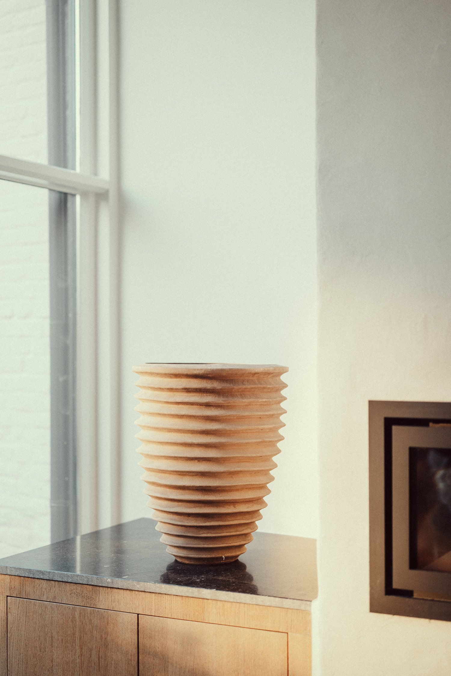 Grote, ribbelige, grijze keramische vaas op een witte vloer naast een witte muur.