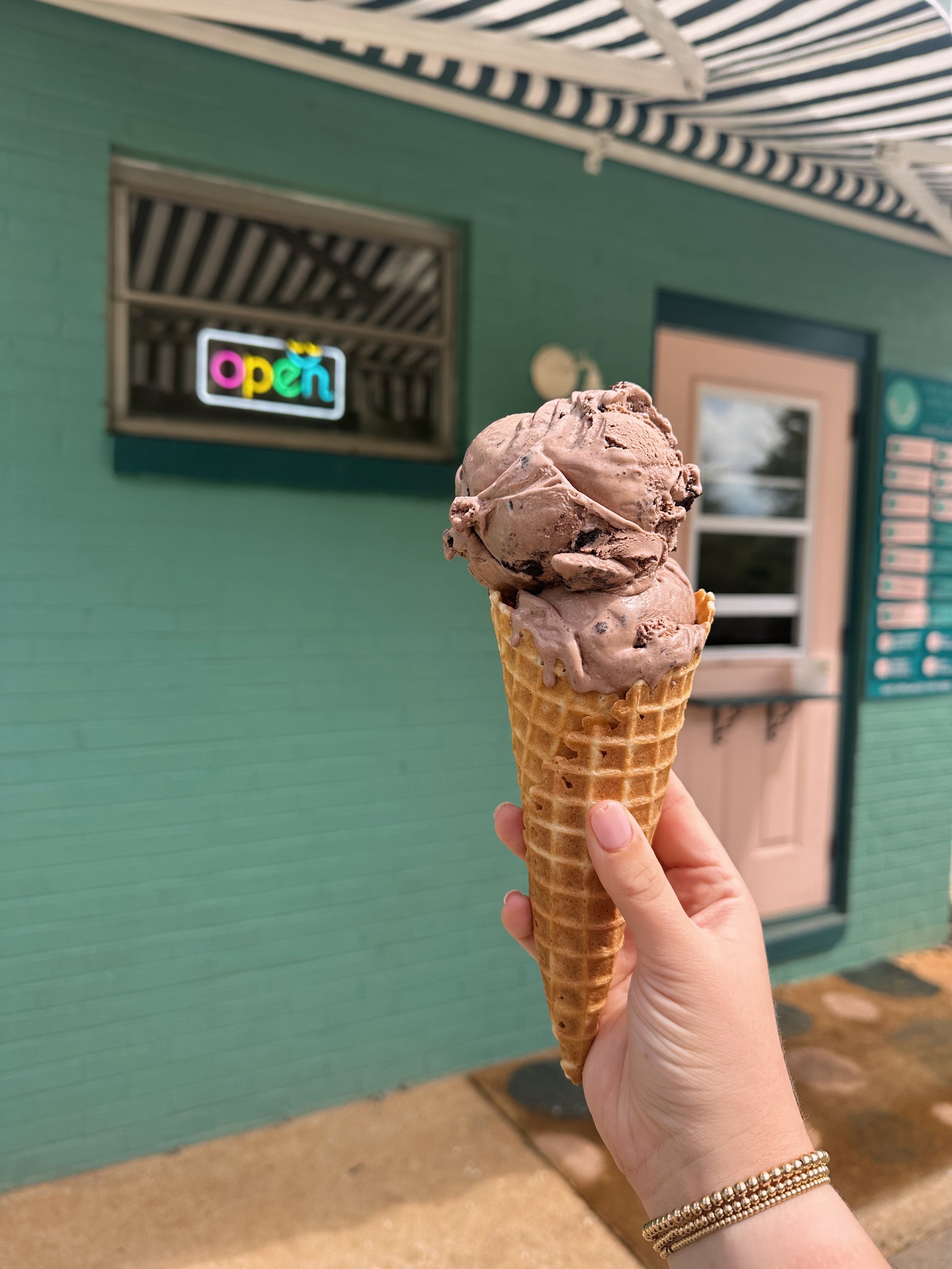 Hand holding a chocolate ice cream in a waffle cone outside a teal building with an "open" sign.