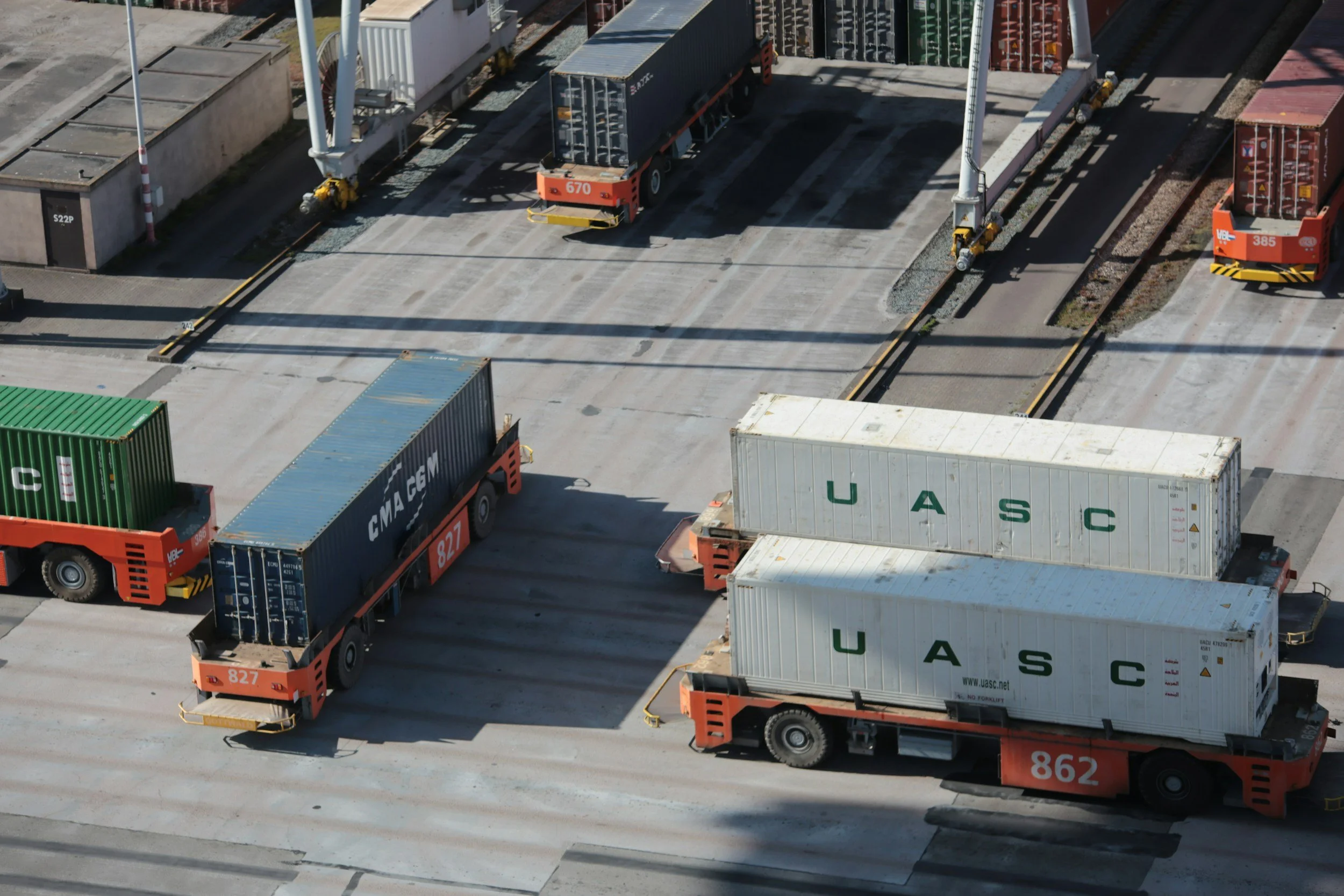 An aerial view of a shipping yard with multiple cargo containers and trucks, some containers are labeled UASC and CMA CGM, with shadow patterns cast by cranes and other equipment.