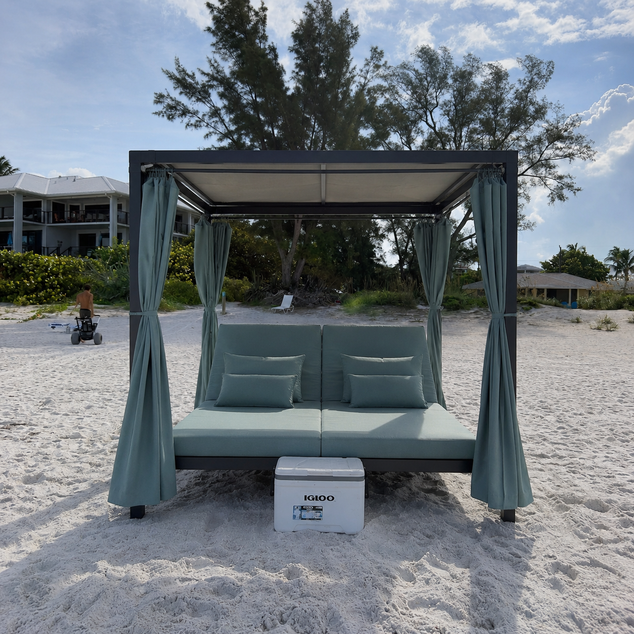 A cushioned canopy bed on the beach, with teal curtains and pillows, an Igloo cooler in front, and a young boy riding a toy vehicle in the background.