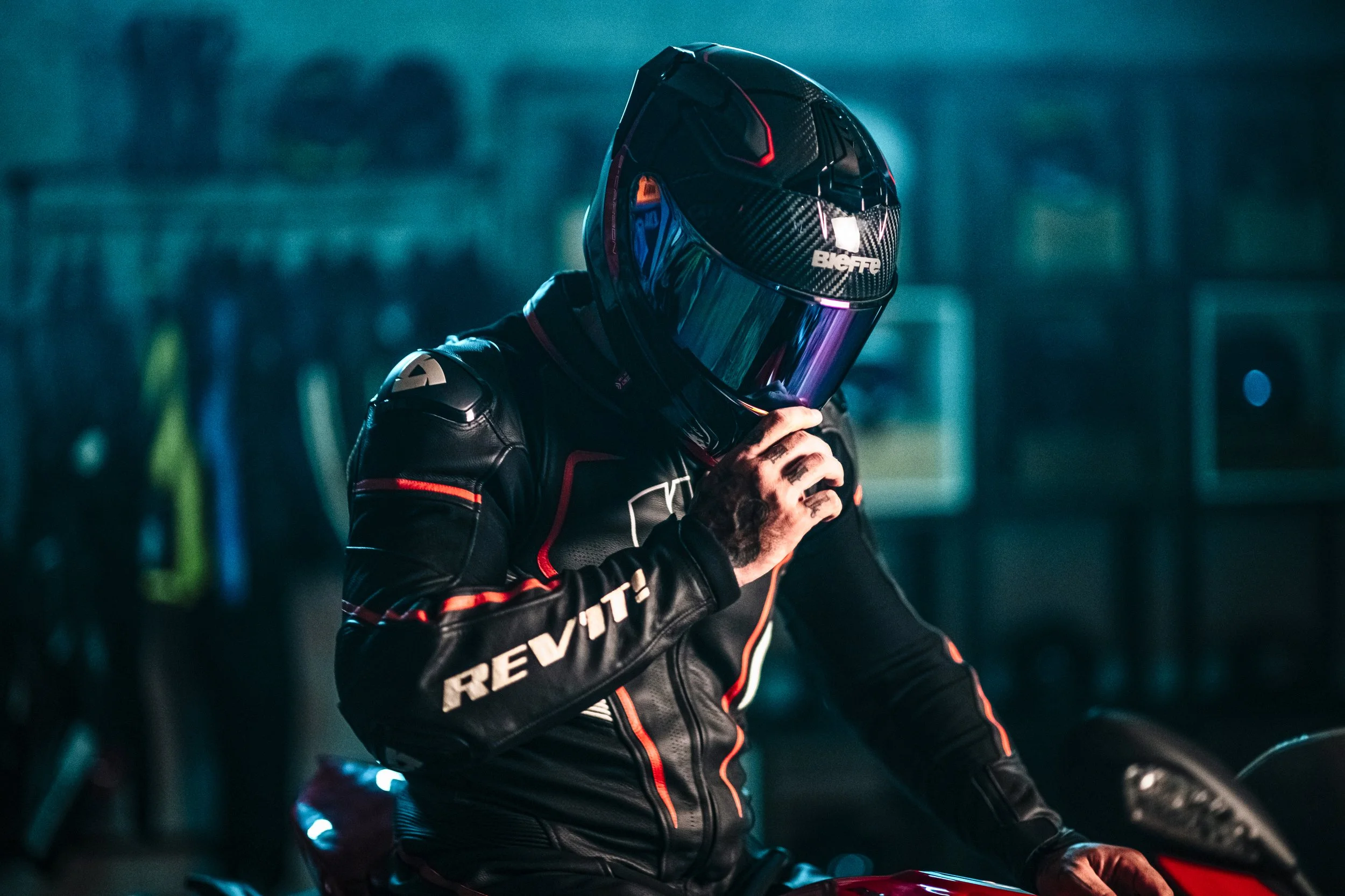 A motorcyclist wearing a black racing suit with red and white accents, a black Bieffe helmet with a reflective visor, preparing for a ride in a dimly lit environment.