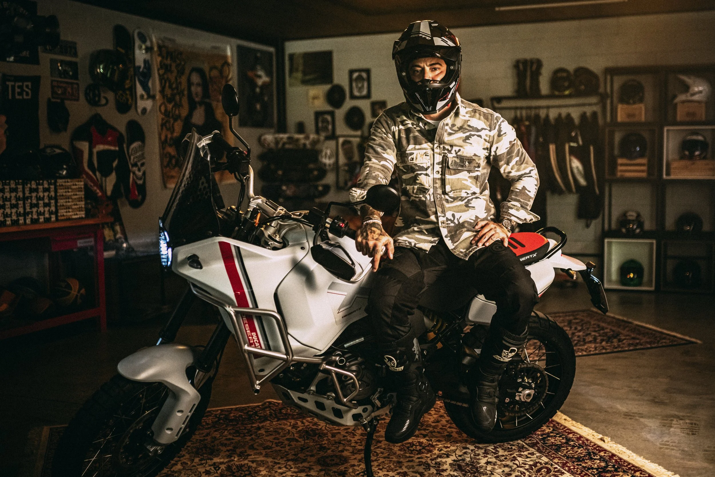 A person wearing a camouflage shirt, black pants, and a full-face helmet, sitting on a white adventure motorcycle inside a room decorated with motorcycle helmets, artwork, and memorabilia.