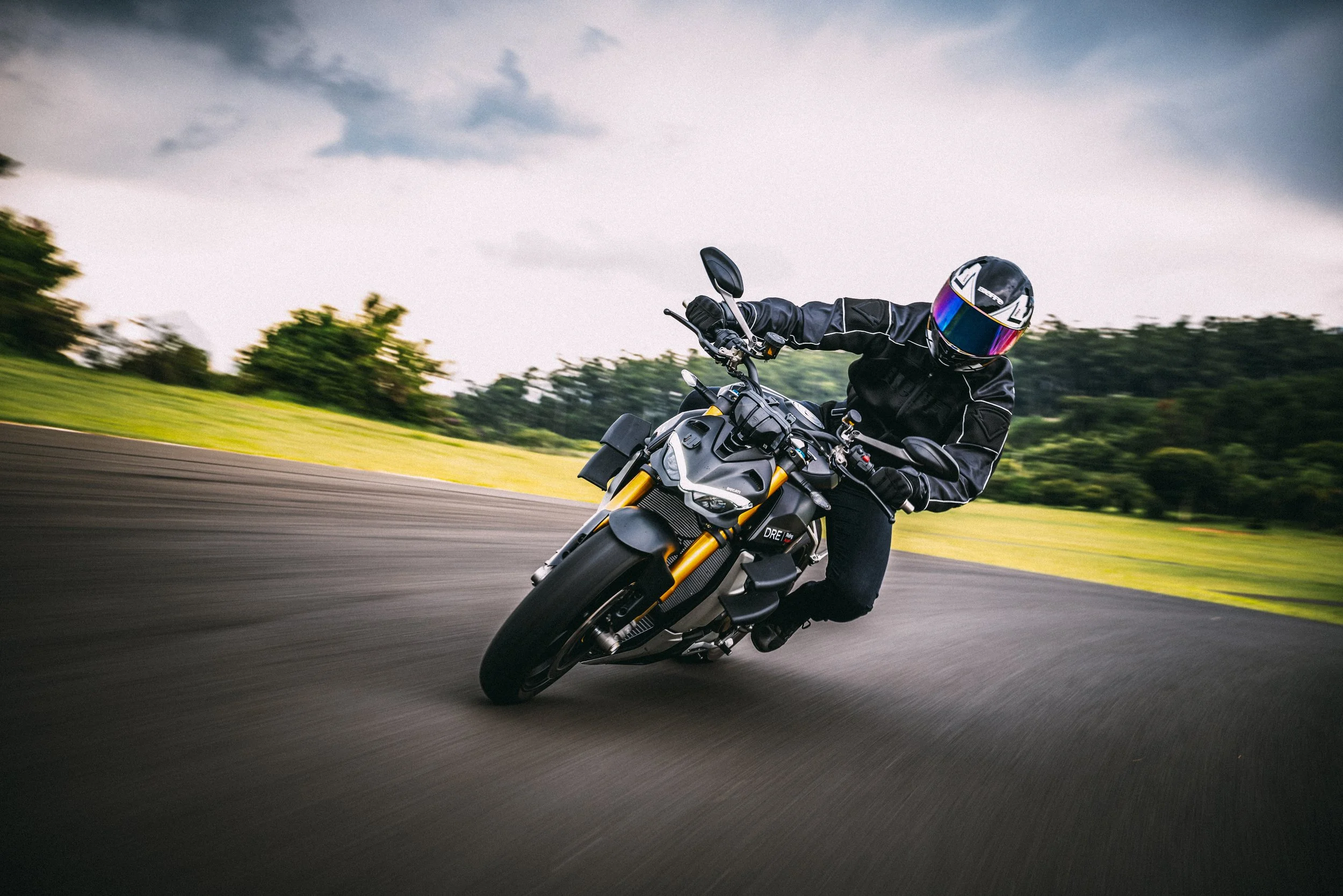 Motorcyclist in black riding gear leaning into a turn on a scenic open road with green fields and trees in the background during daytime.