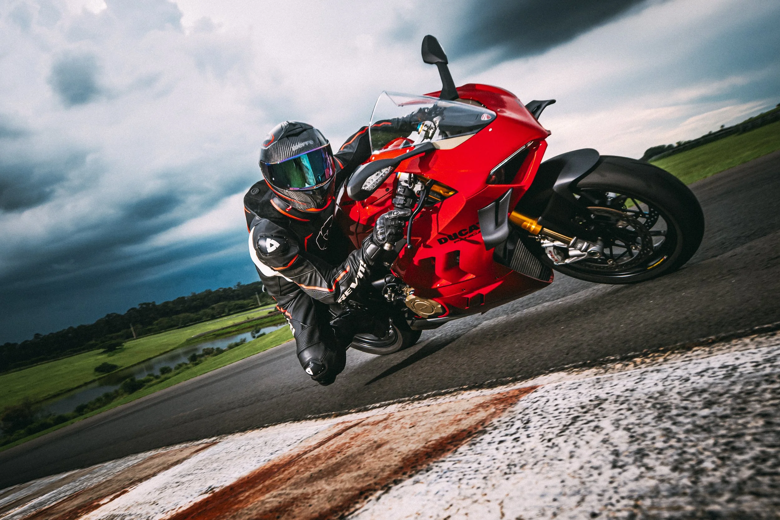 Motorcycle racer wearing black helmet and suit leaning into a turn on a racetrack with green fields and cloudy sky in the background.