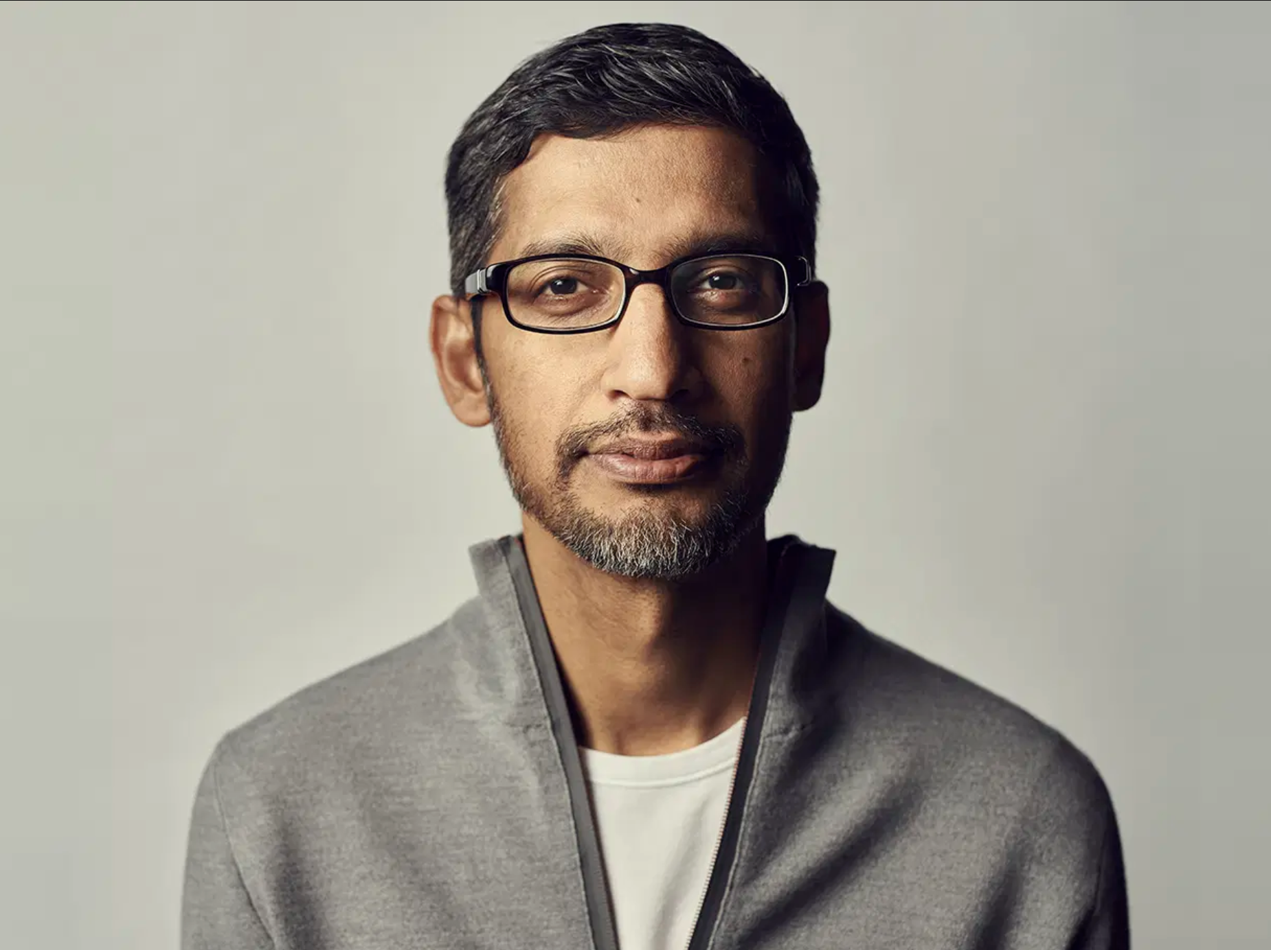 Sundar Pichai wearing glasses and a gray jacket against a neutral background.