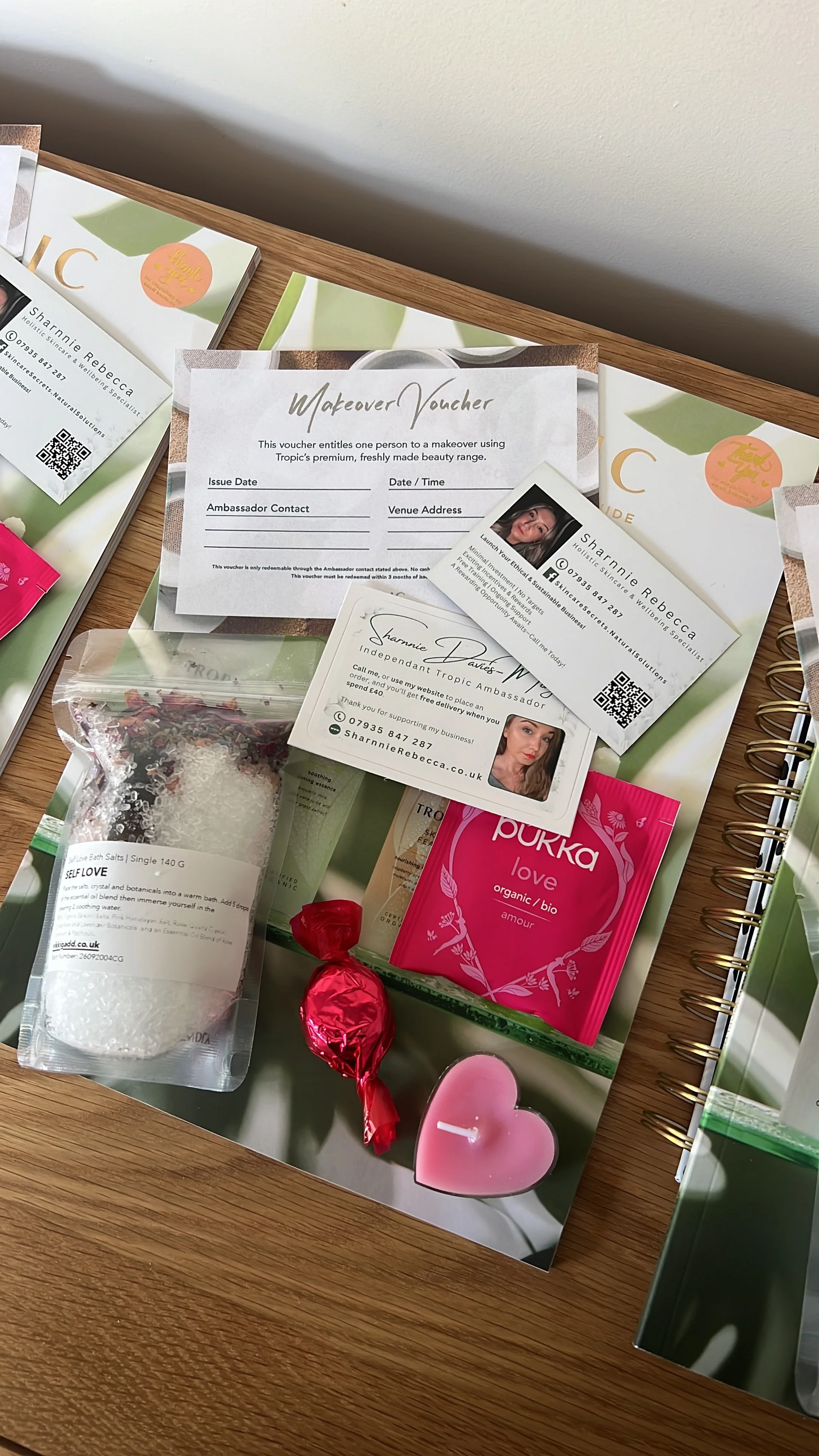 A table filled with promotional materials and gifts, including a makeover voucher, business cards, bath salts, a pink wrapped candy, a pink candle, and a pink greeting card with a heart. The setup appears to be for a beauty or wellness event.