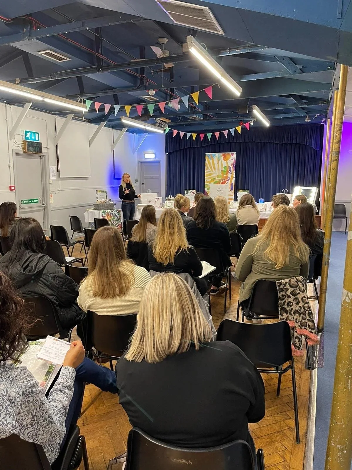 A woman standing on a stage with a microphone, speaking to an audience seated in a room decorated with colorful bunting. The audience appears engaged, with some taking notes or looking at the speaker. The room has a dark ceiling with exposed beams, bright lighting, and a blue curtain backdrop.