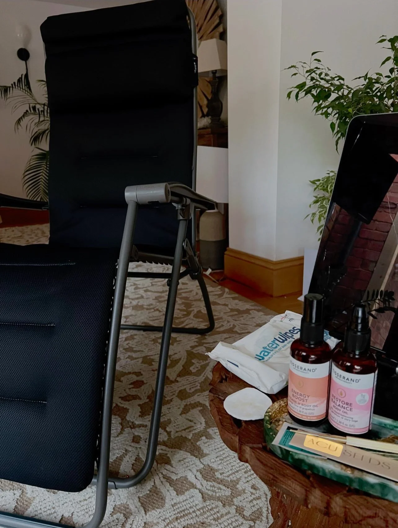 Hotel room with a black reclining chair, a side table with bottles of essential oil, a pile of wipes, and a laptop, with plants and a patterned carpet in the background.
