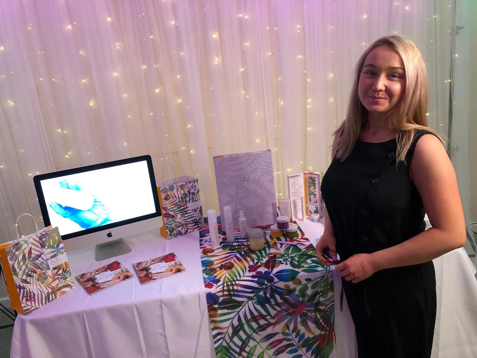 A woman standing next to a table with skincare products, gift bags, and promotional materials, with a backdrop of string lights.
