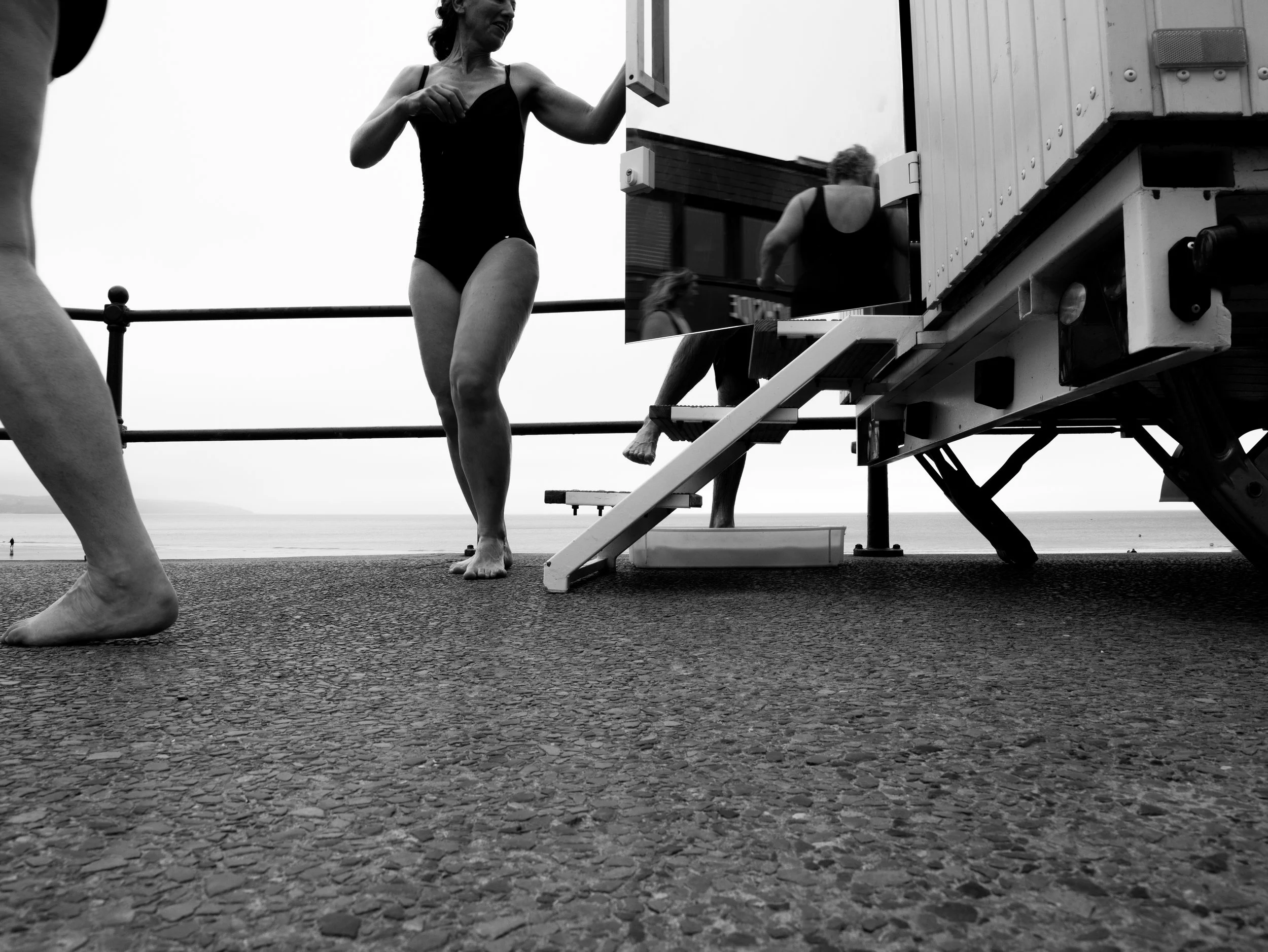Black and white photo of women in swimsuits at the beach, with a lifeguard tower or structure in the background and a view of the ocean.