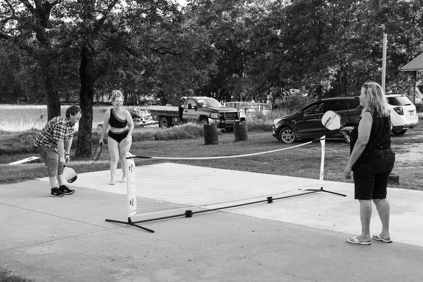 Three people playing a game of pickleball outdoors on a concrete court, with cars and trees in the background.