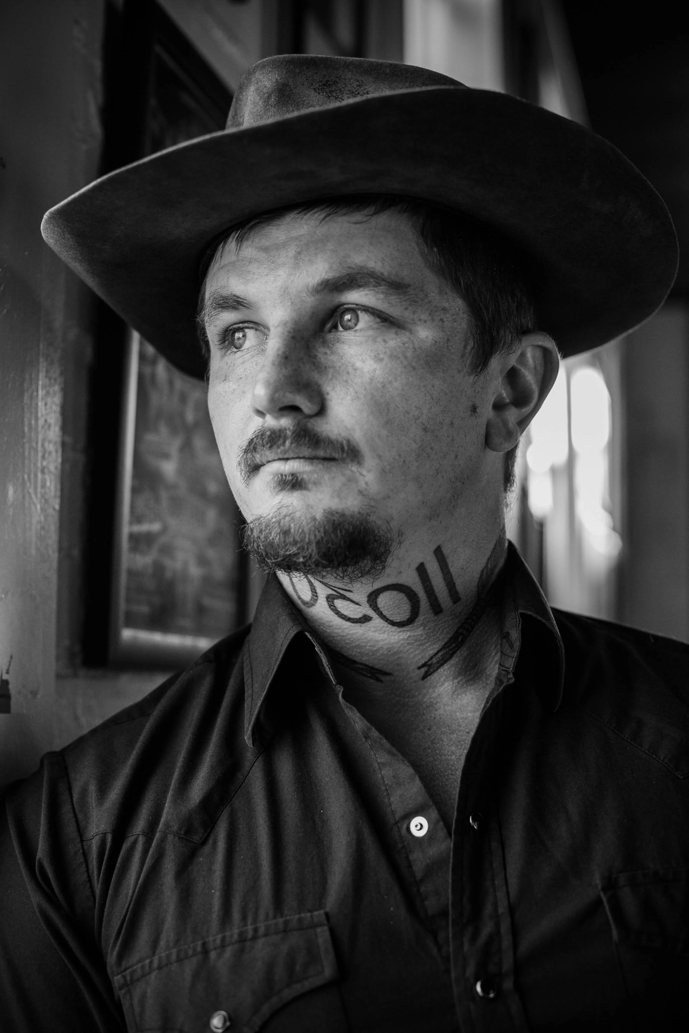 A black-and-white portrait of a young man wearing a cowboy hat, with visible tattoos on his neck, and a goatee. He is looking to the side, with light coming from the right.