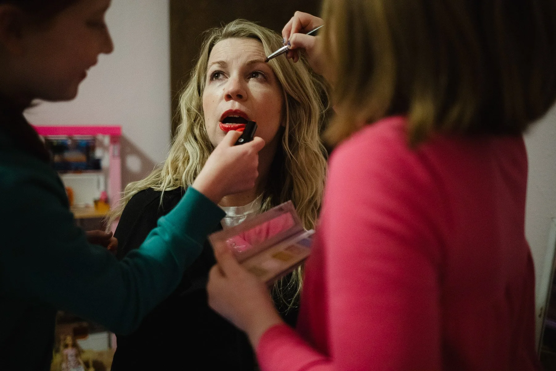 A woman with blonde hair is getting her makeup done by two makeup artists; one is applying lipstick, and the other is doing her eyeshadow or eyebrow.