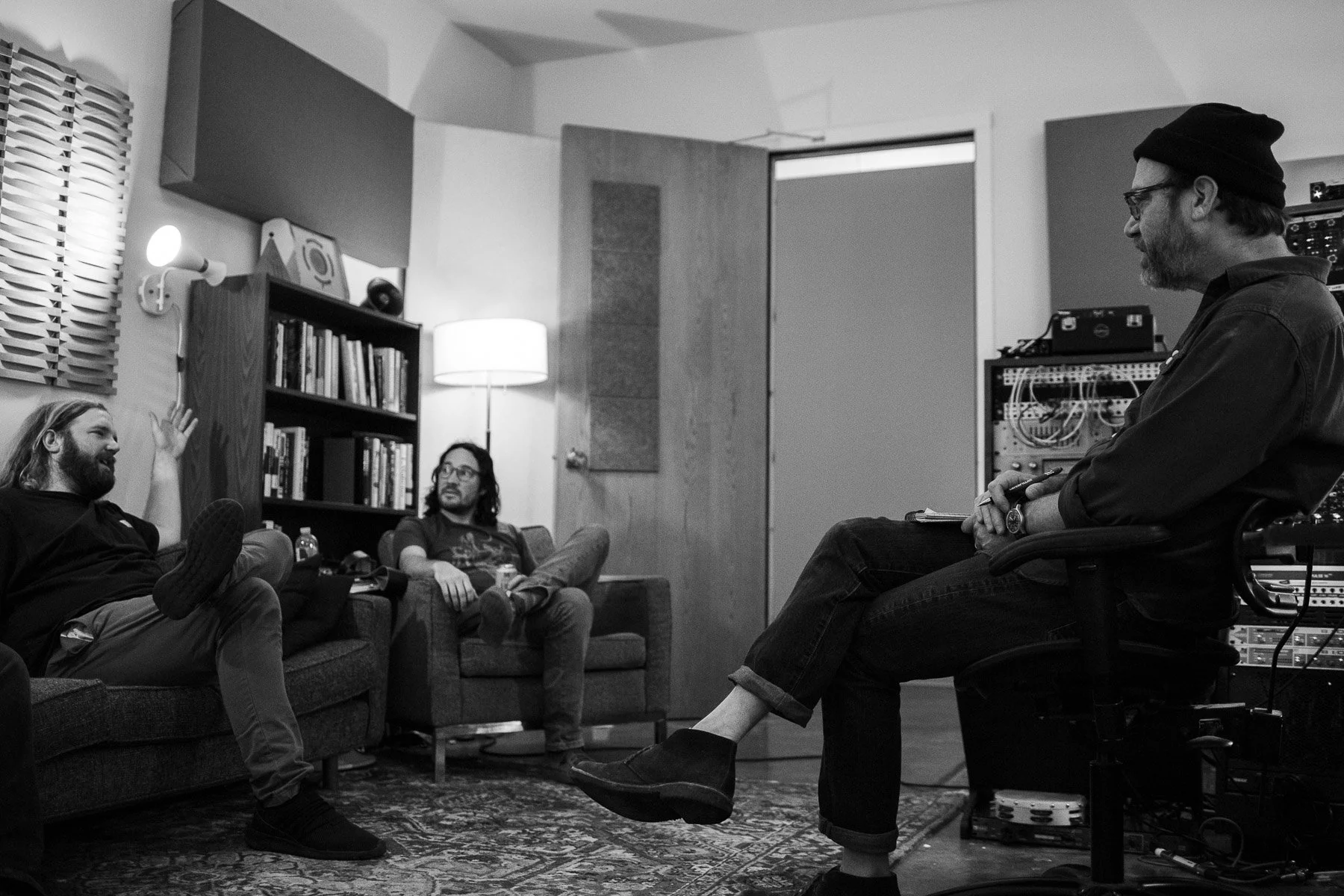 Three men in a recording studio having a discussion. Two men sit on a sofa, one raises his hand and talks, while the third man sits on an office chair with headphones on, holding a pen. There are bookshelves and studio equipment in the background, an