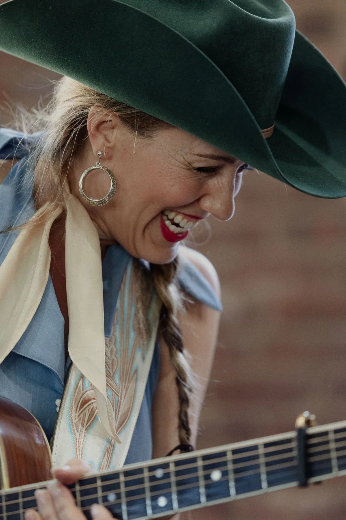 A woman wearing a large green hat, red lipstick, and hoop earrings, smiling while playing a guitar.