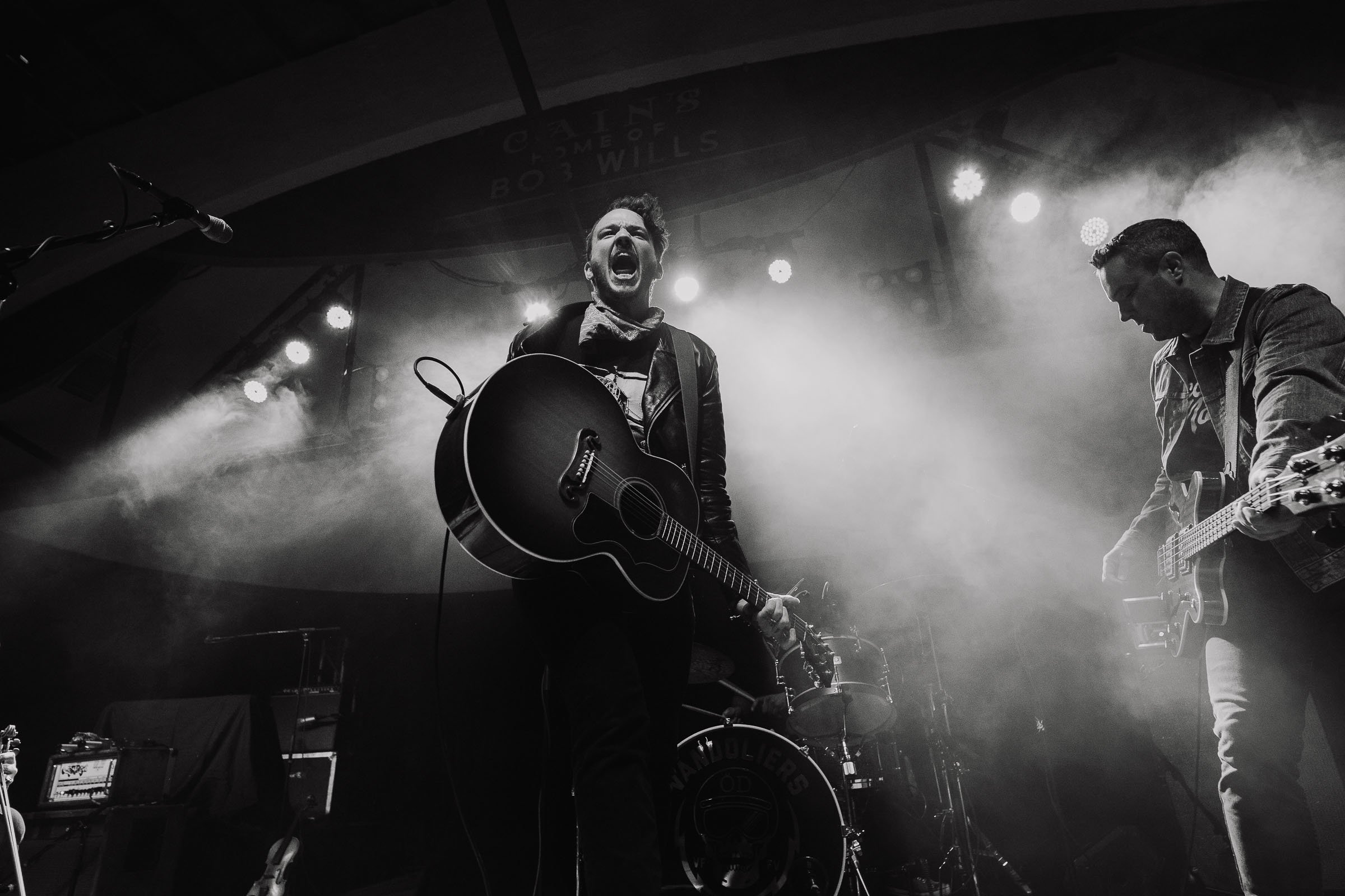 Black and white photo of a band performing on stage, with one singer playing an acoustic guitar and singing passionately, and members playing electric guitars in the background, stage lights creating a smoky atmosphere.