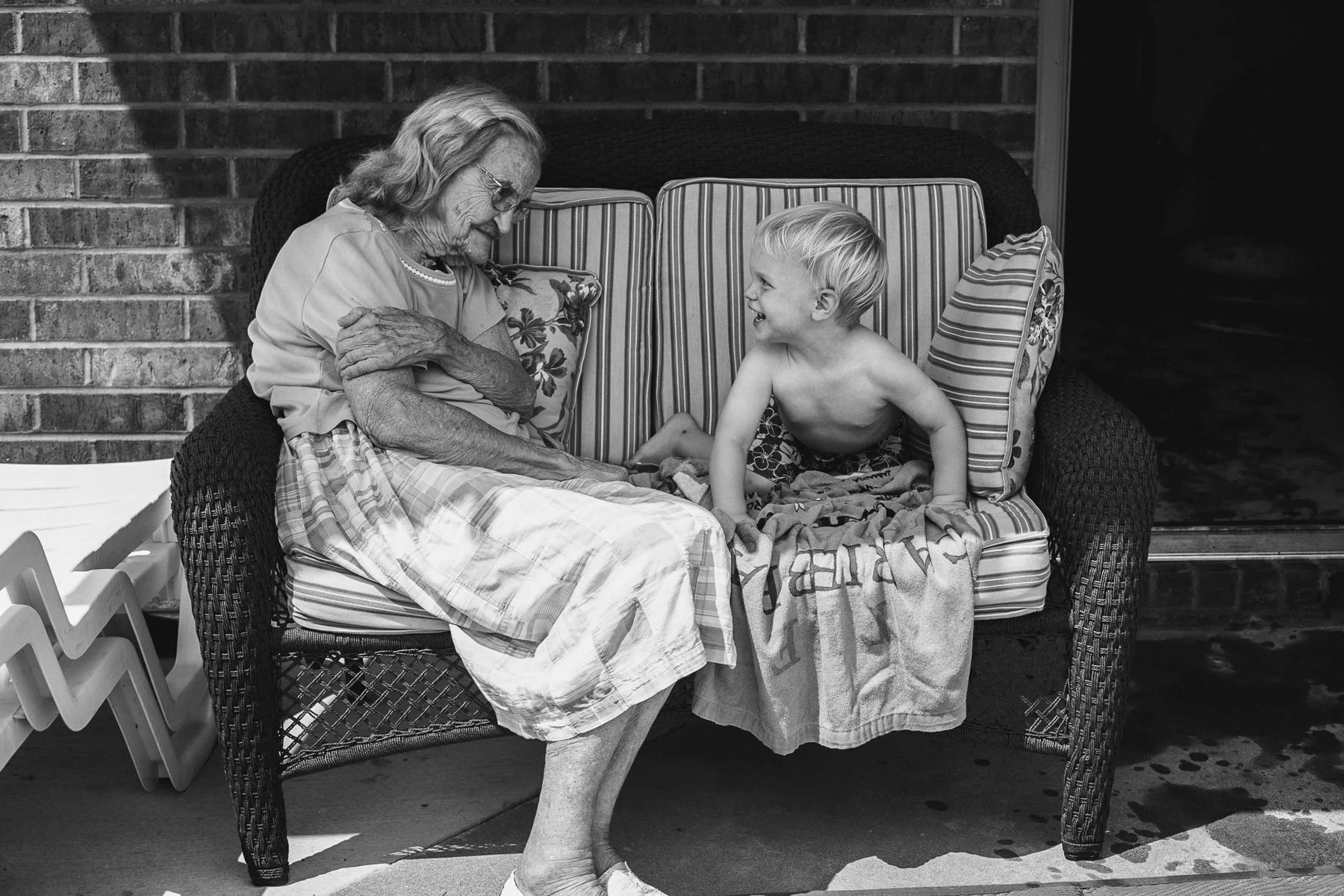 A smiling young boy and an elderly woman sit on a wicker couch, sharing a joyful moment outdoors. The woman is wearing glasses and a blouse, while the boy is shirtless and wearing shorts. They are engaged in a cheerful conversation, with pillows and 