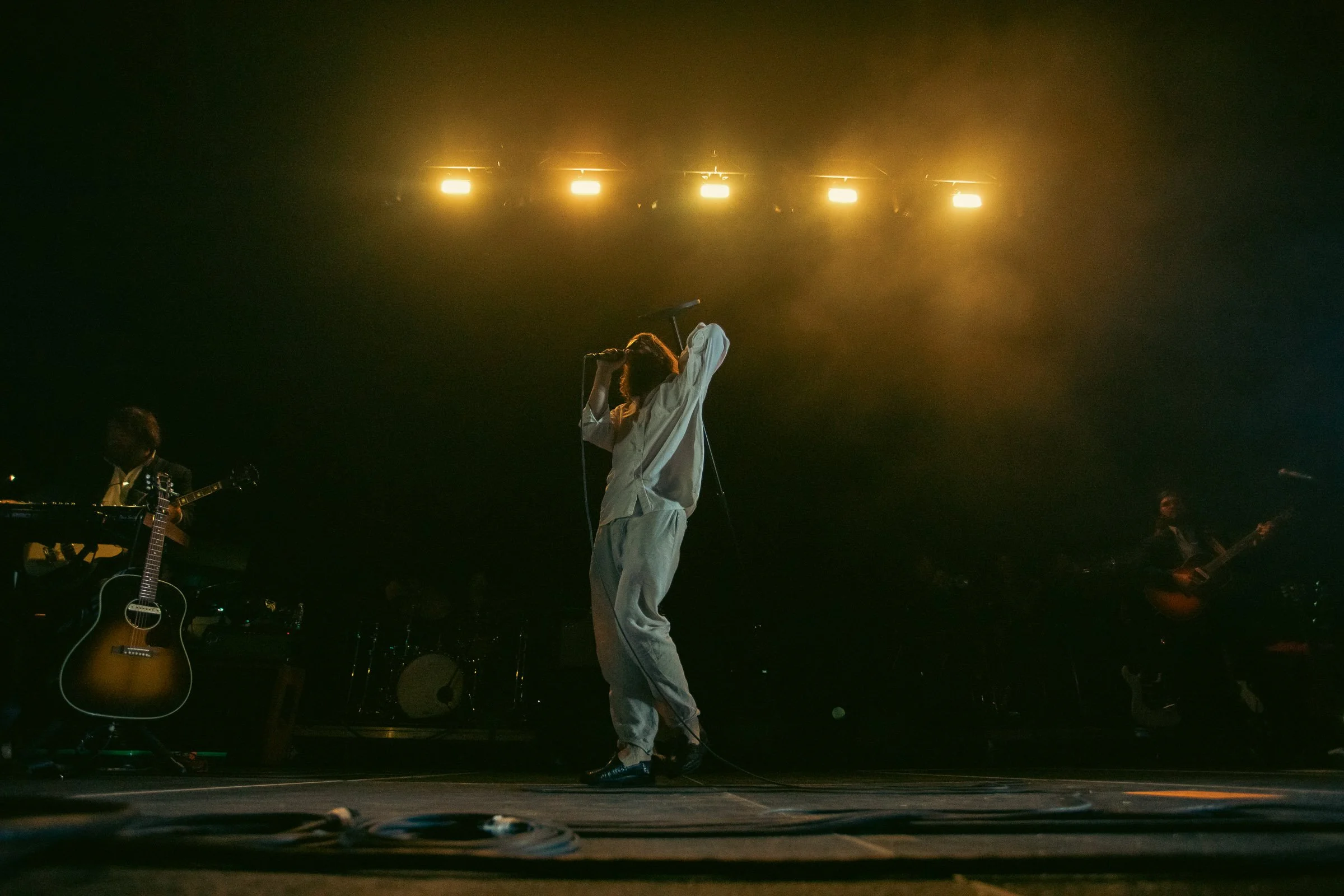 Musician performing on stage with backup band, holding microphone and wearing loose clothing, under yellow stage lights.