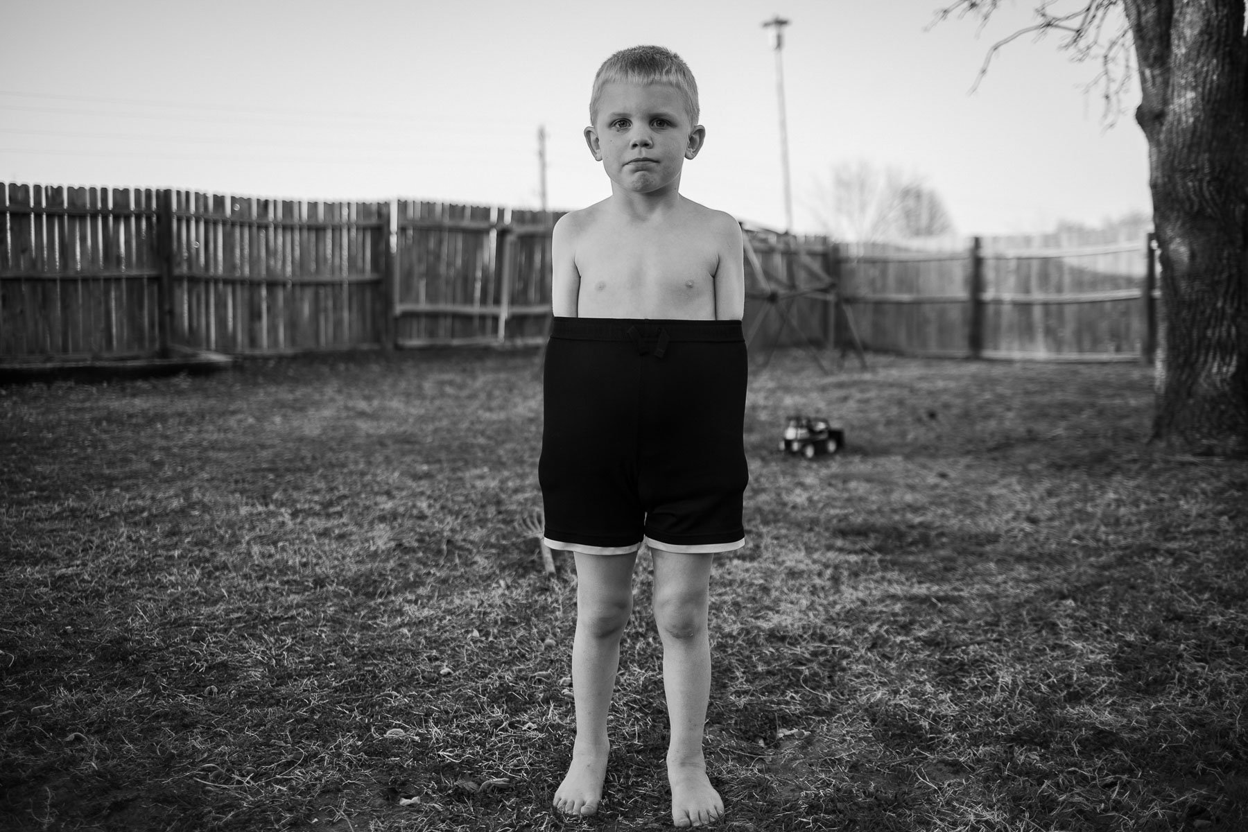 Black and white photo of a young boy standing barefoot on grass in a backyard, with a wooden fence, a tree, and some outdoor toys in the background.