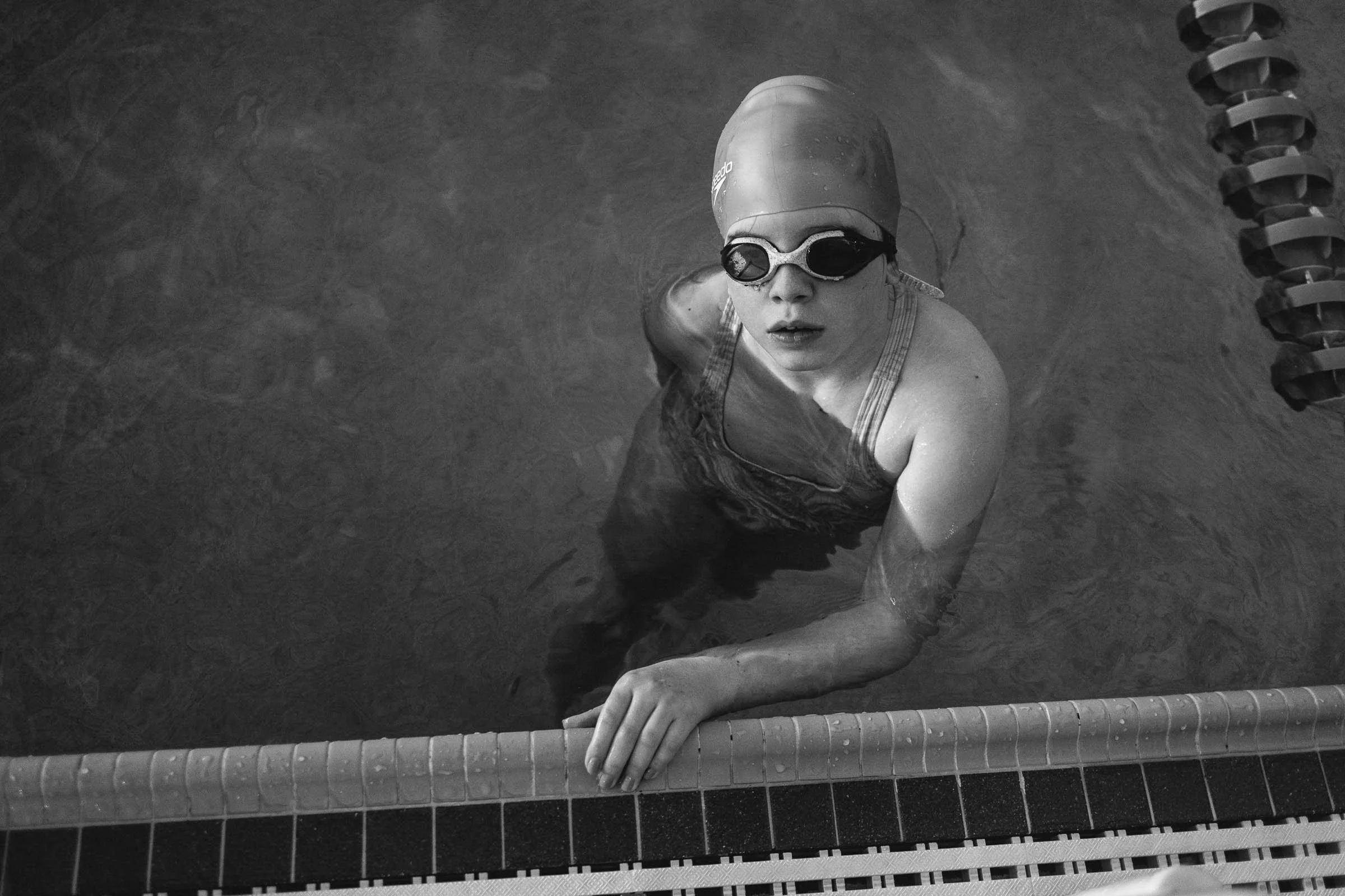 A young woman in swimming goggles and a swim cap, holding onto the pool edge, looking upward in a swimming pool.