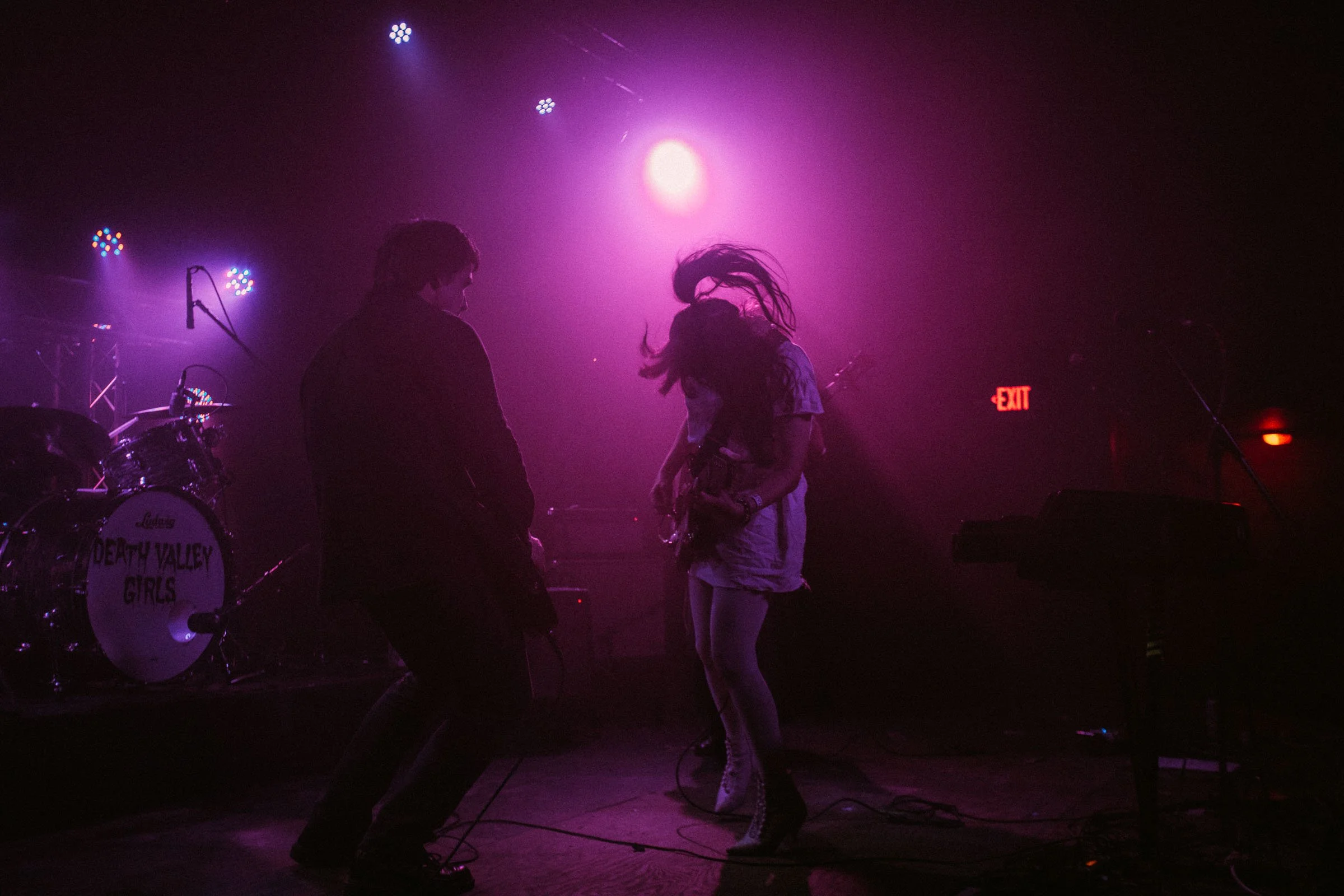 Silhouettes of two musicians performing on stage with purple and pink lighting, with a drum set labeled 'Death Valley Girls' in the background and an illuminated exit sign.
