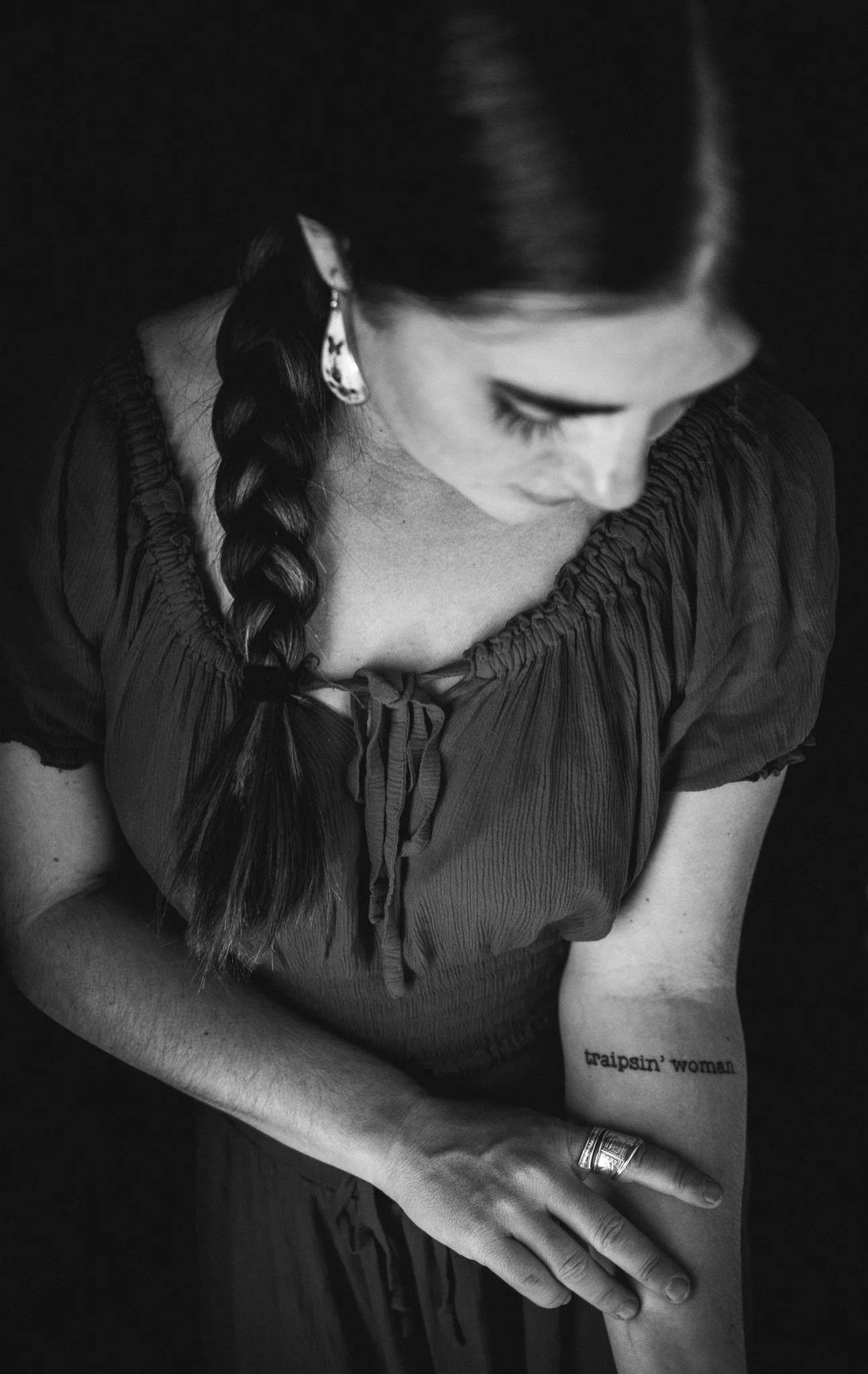 Black and white photo of a woman with braided hair, wearing earrings and a dark top, looking down. She has a tattoo on her forearm that reads 'traipsin' woman' and is wearing a ring on her finger.