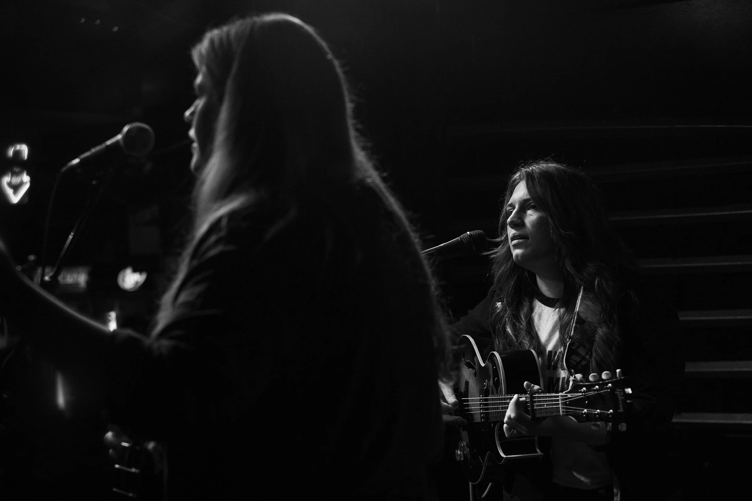 Two women playing guitars and singing into microphones during a performance in a dark setting, with one woman's face visible in profile and the other's face partially visible.