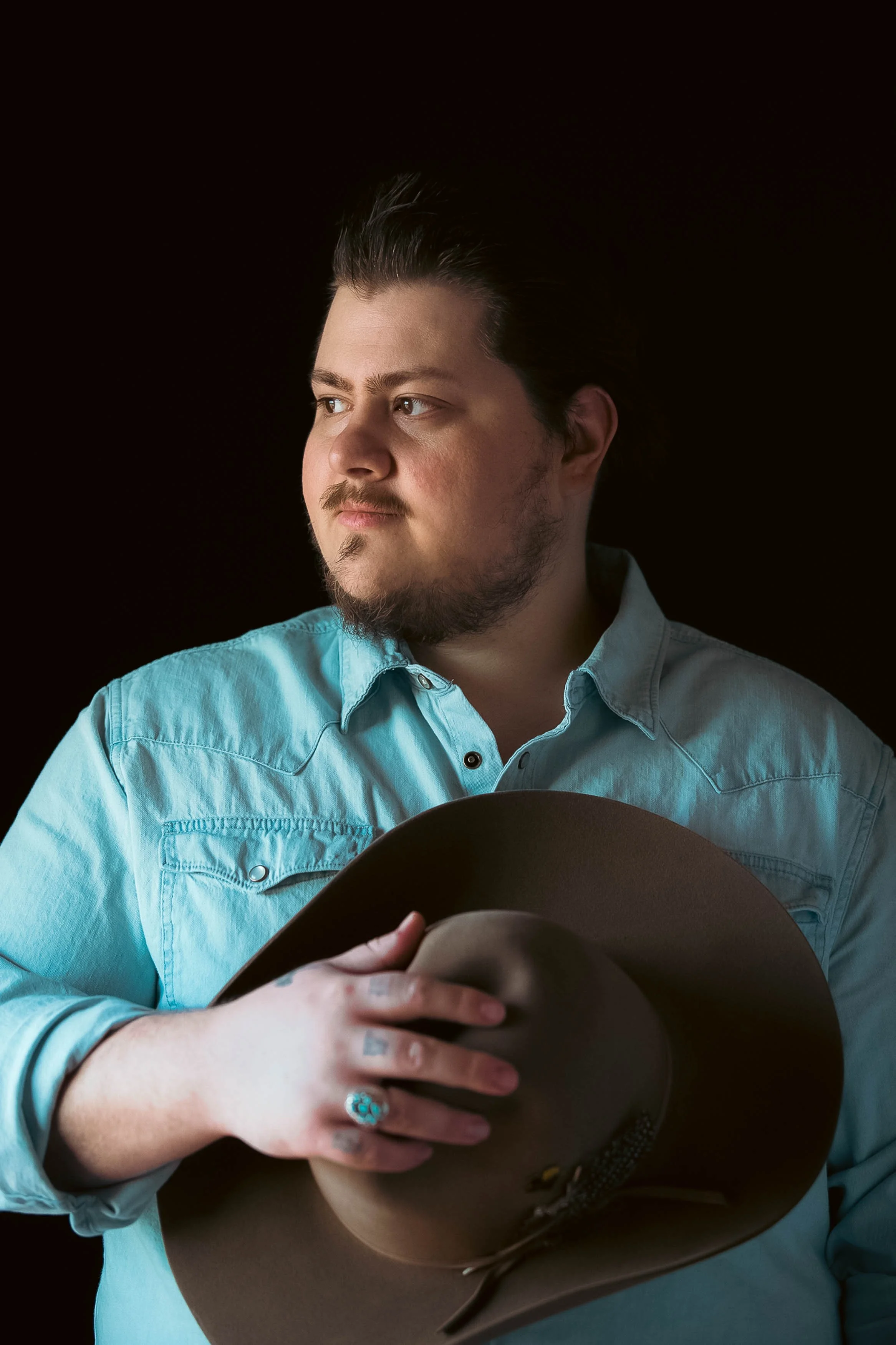 Man with dark hair, beard, and mustache wearing a light blue shirt, holding a brown cowboy hat, looking to the side against a black background.