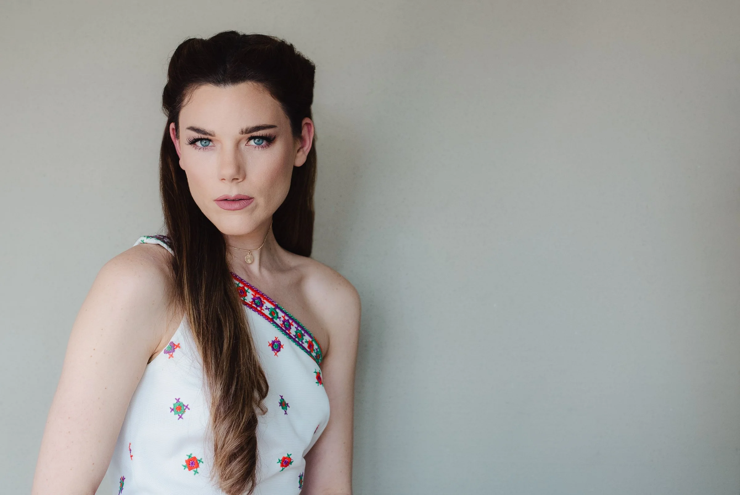 A woman with long dark brown hair and bright blue eyes standing against a neutral gray background, wearing a sleeveless white top with colorful embroidered pattern.