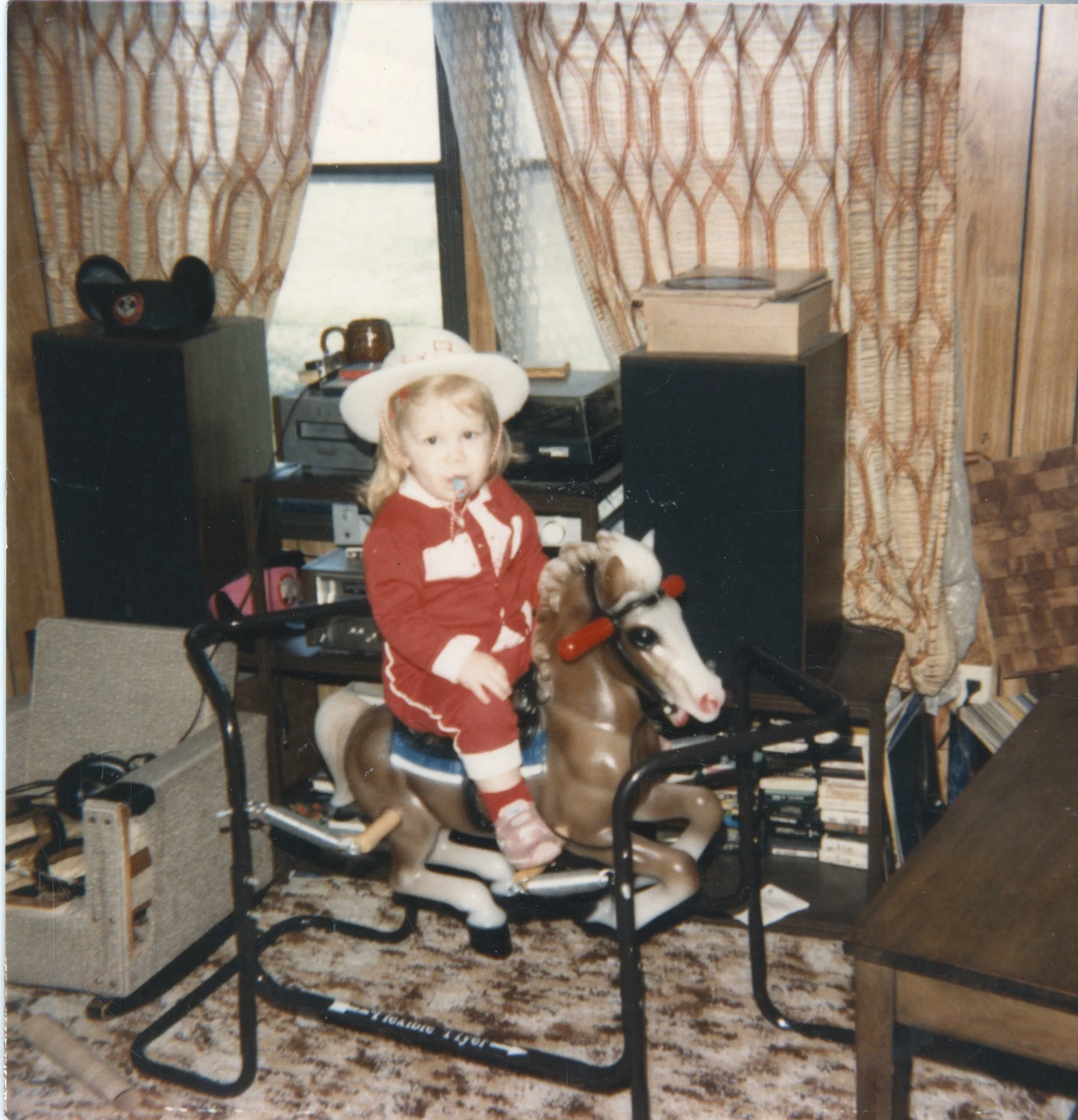 A young girl dressed in a red and white outfit with a white hat, riding a vintage rocking horse in a cozy living room with wood-paneled walls, heavy patterned curtains, and various furniture and electronics around.