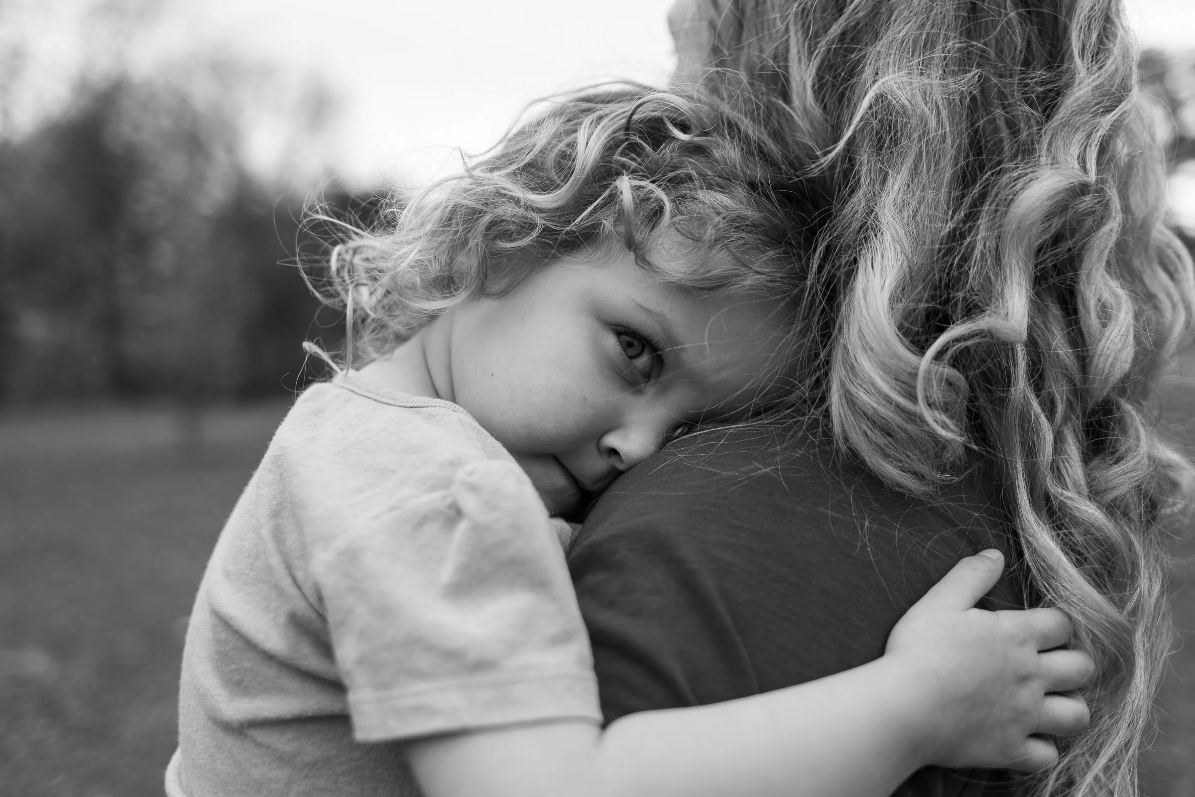 A young girl with curly hair hugging an adult with curly hair in an outdoor setting.