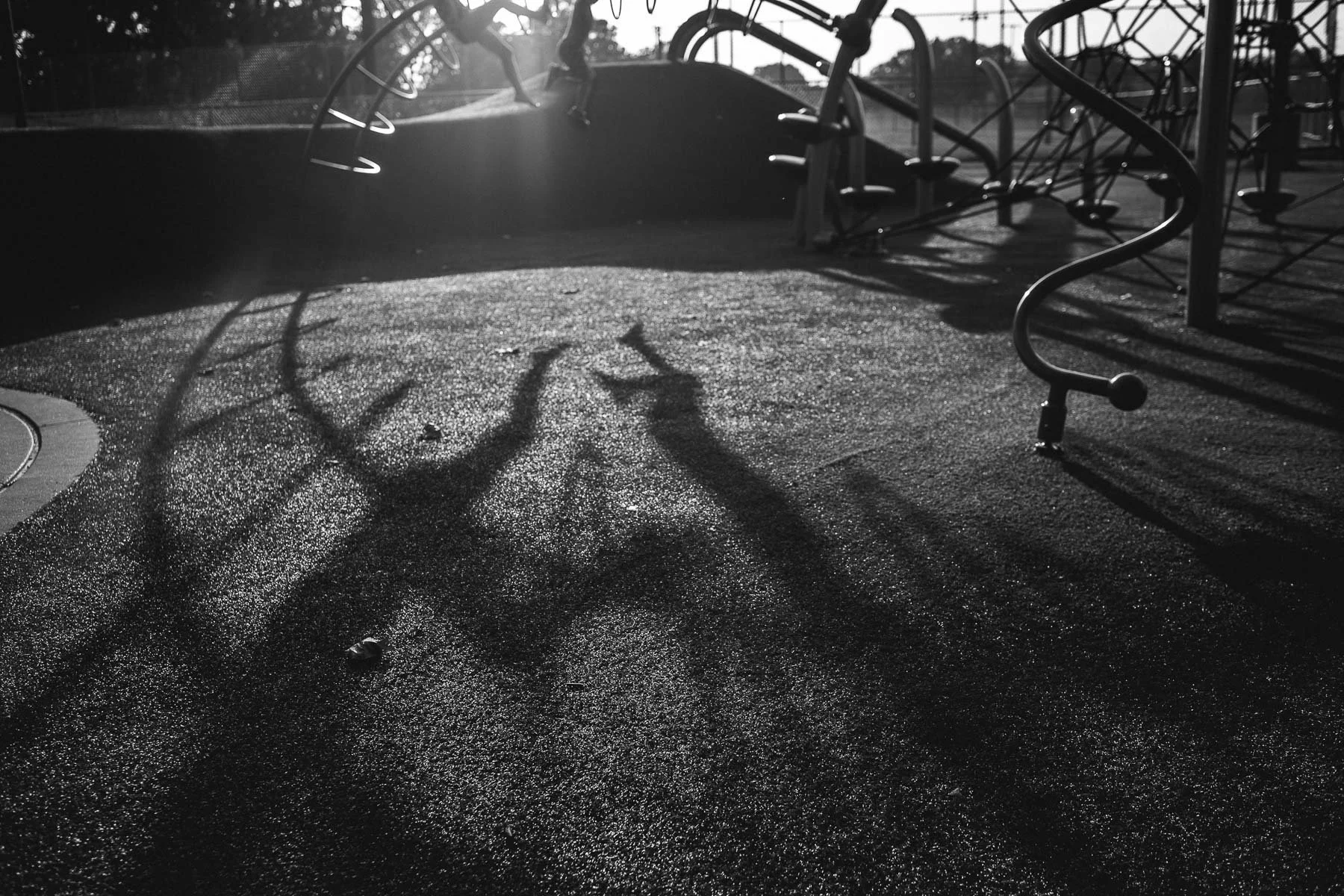 A playground with metal climbing structures, cast long shadows on a wet, textured ground, illuminated by sunlight.