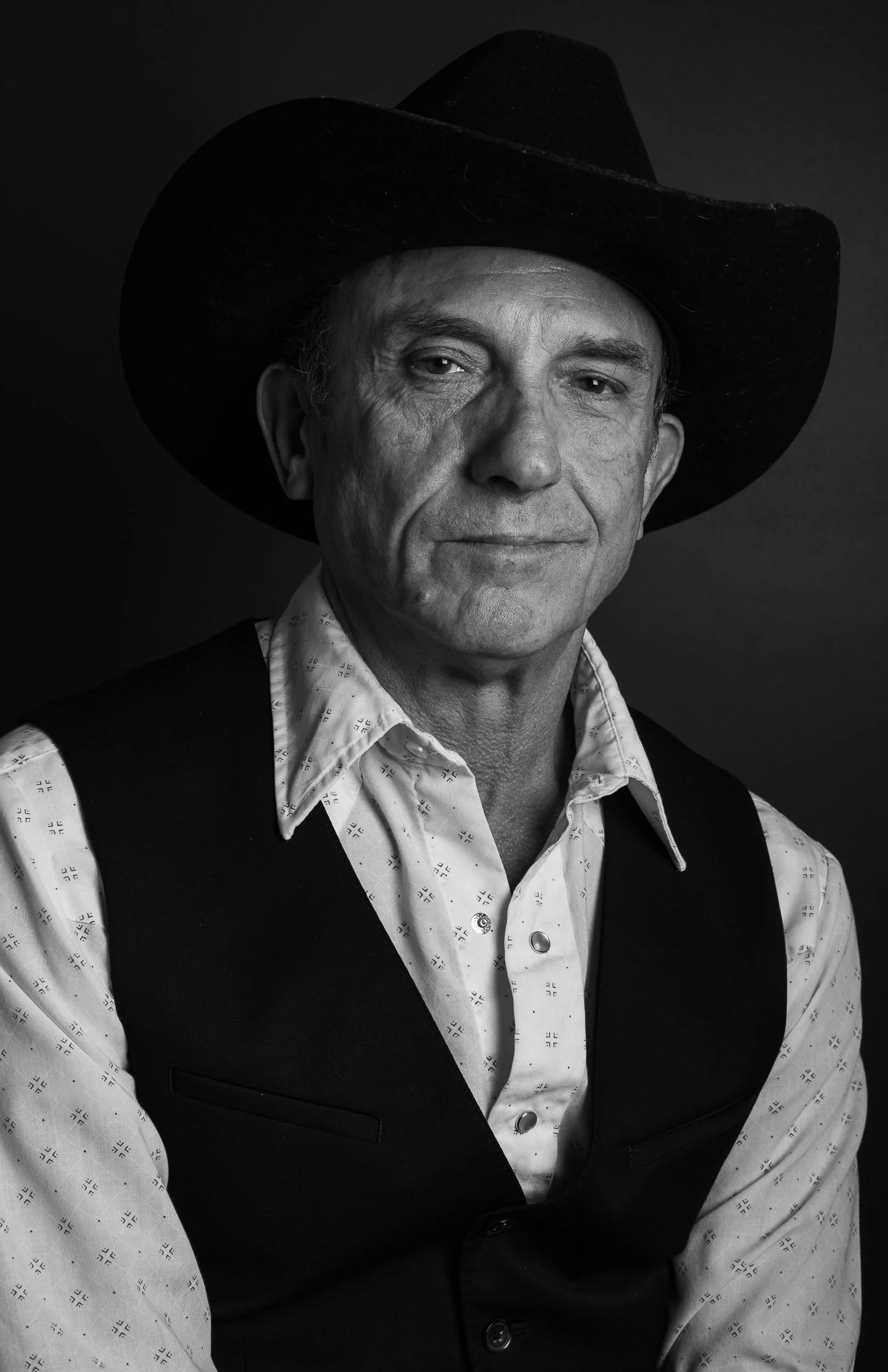 Black and white portrait of a middle-aged man wearing a cowboy hat, a patterned white shirt, and a dark vest. He has a serious expression and is looking directly at the camera.