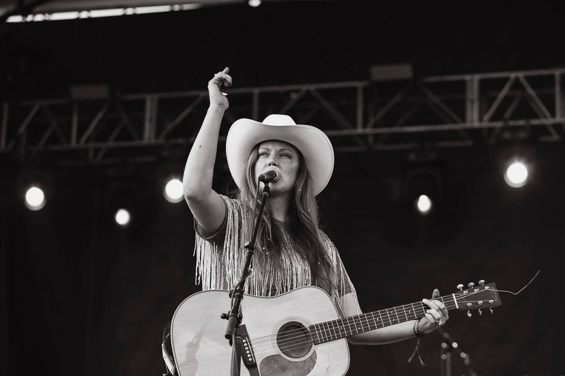 A woman performing on stage with a guitar, wearing a white cowboy hat and a fringed top, singing into a microphone.
