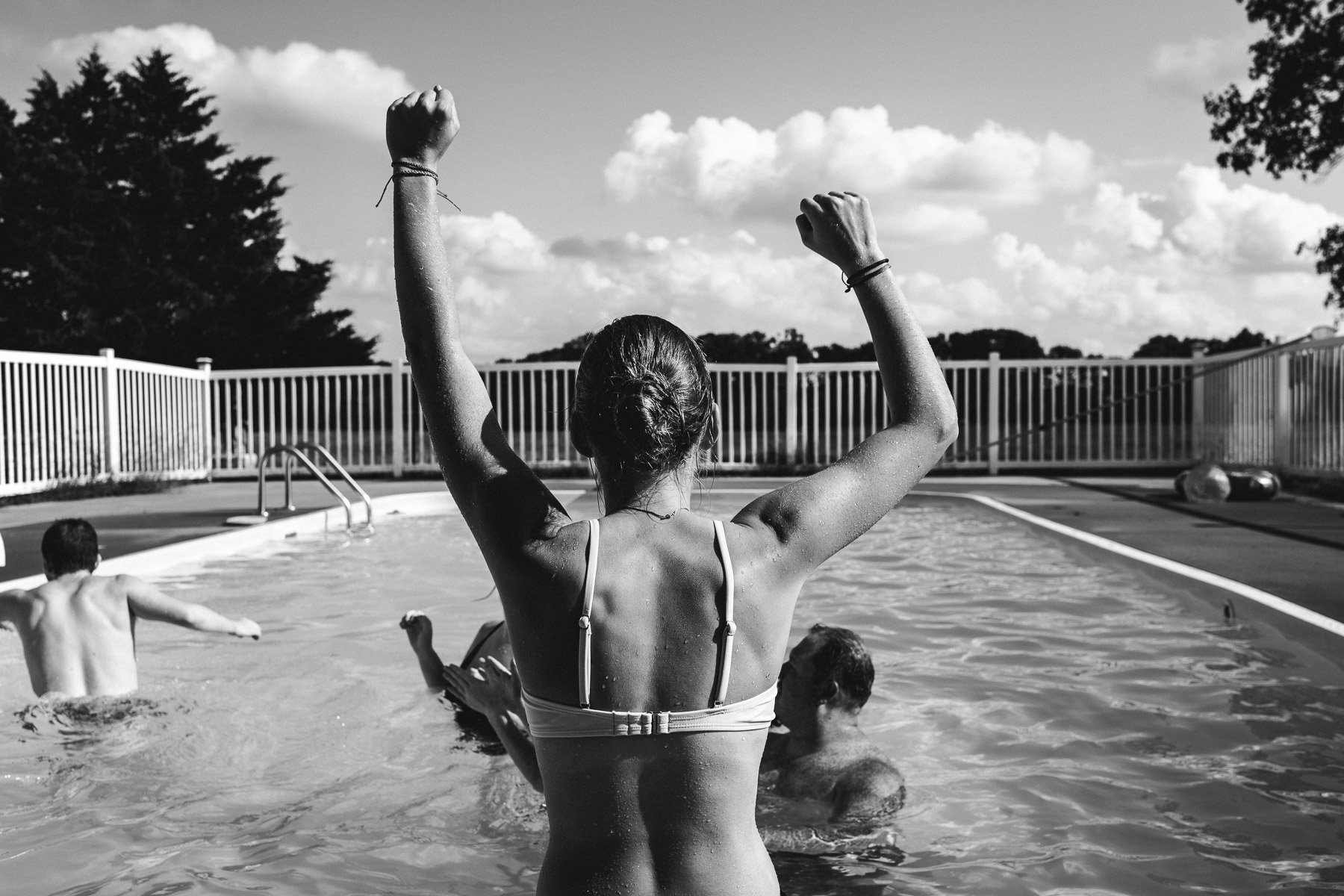 A woman in a swimsuit raising her arms in a pool with children swimming around her. Black and white image.