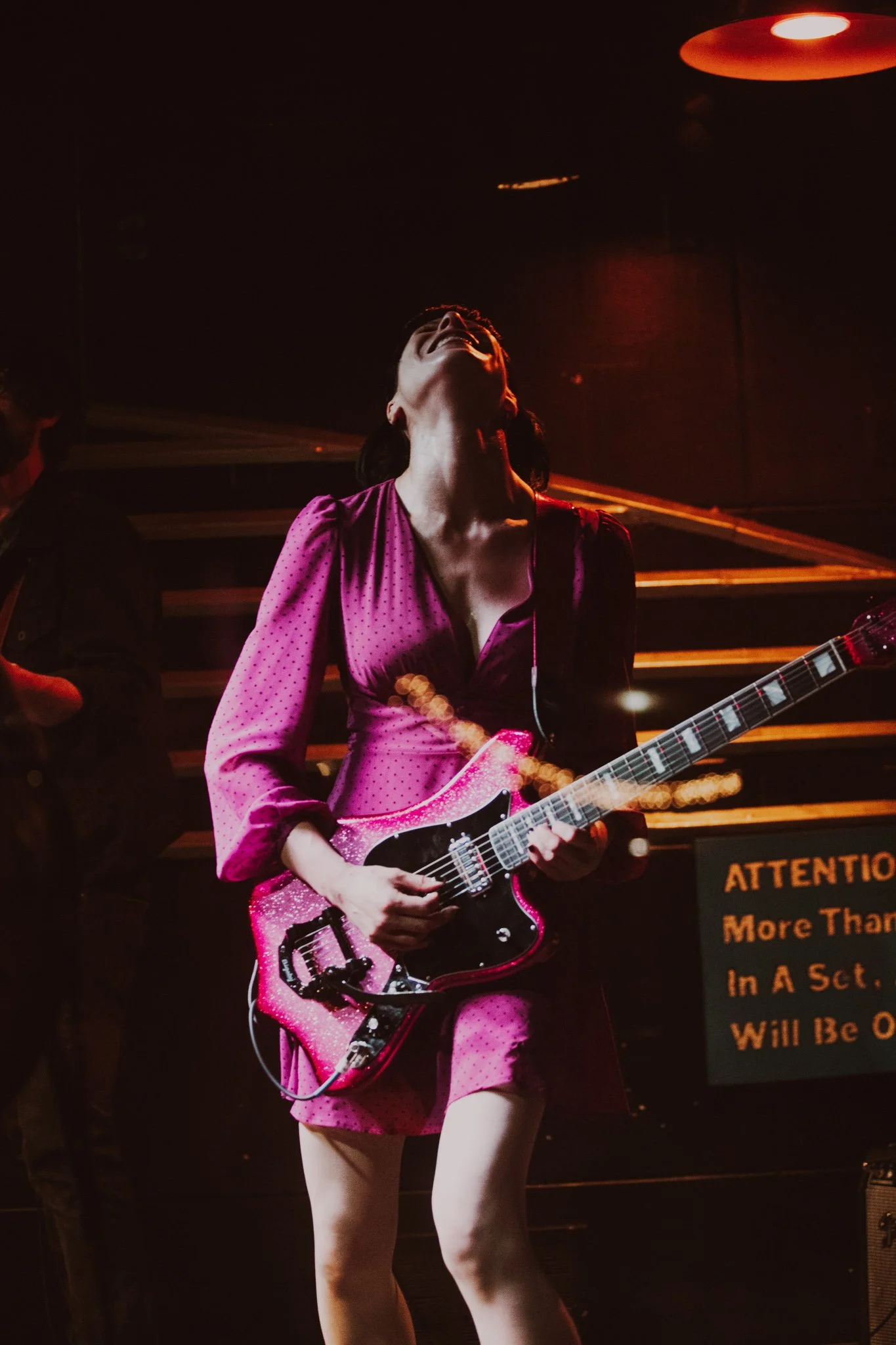 A woman wearing a pink dress playing an electric guitar on stage, with a dark background and stage lights.
