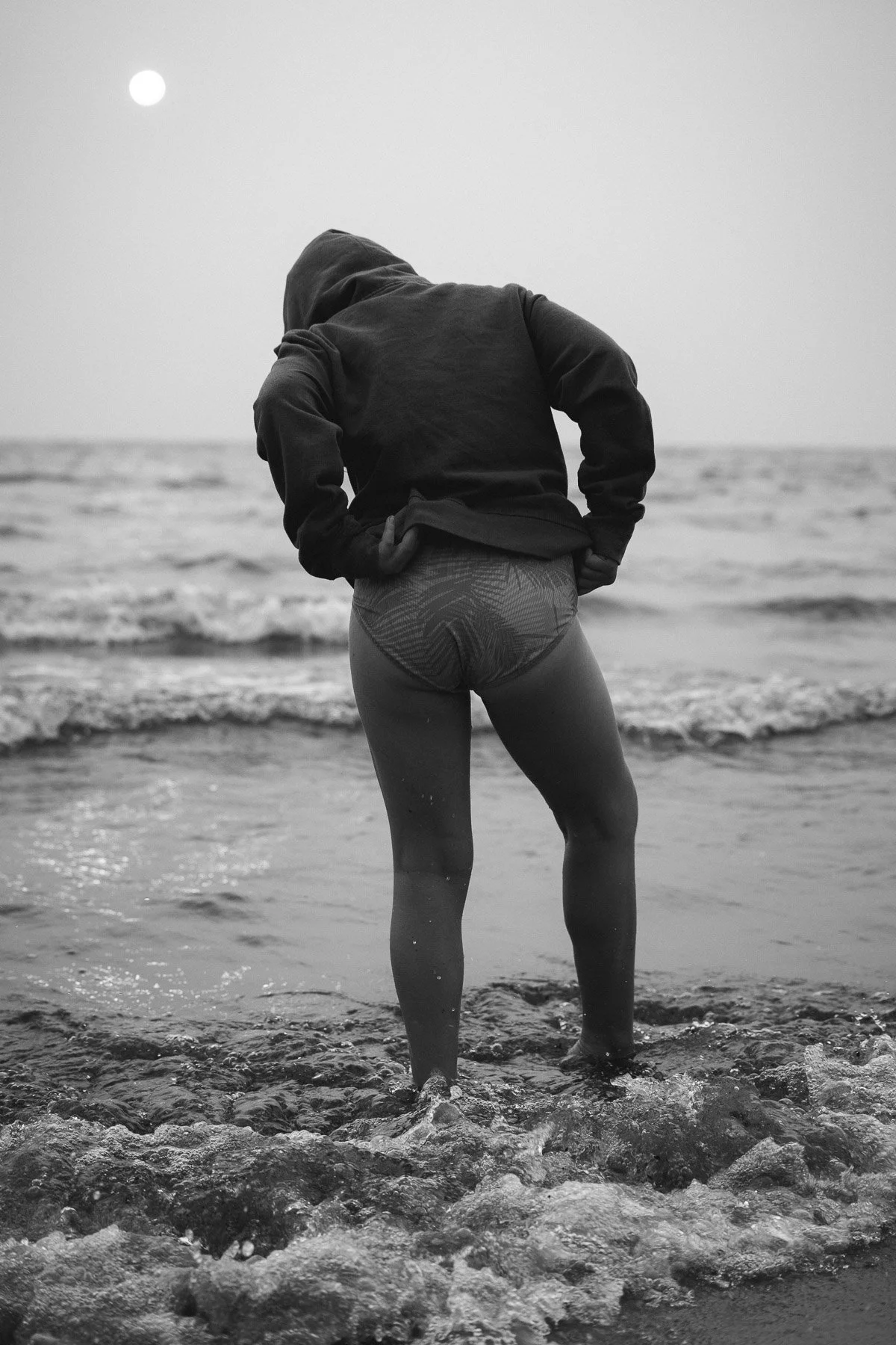 A person standing in the ocean with their back to the camera, wearing a hoodie and patterned shorts, looking out at the water with the sun in the sky.