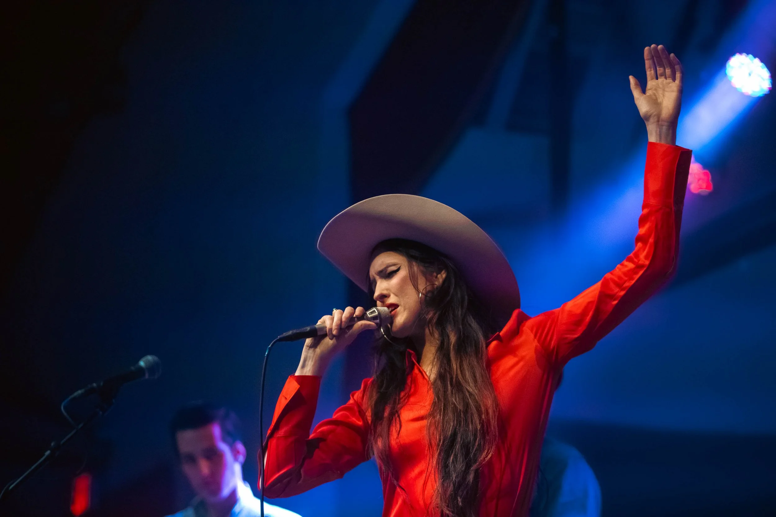 A woman in a red dress and large hat sings into a microphone on stage with blue lighting.