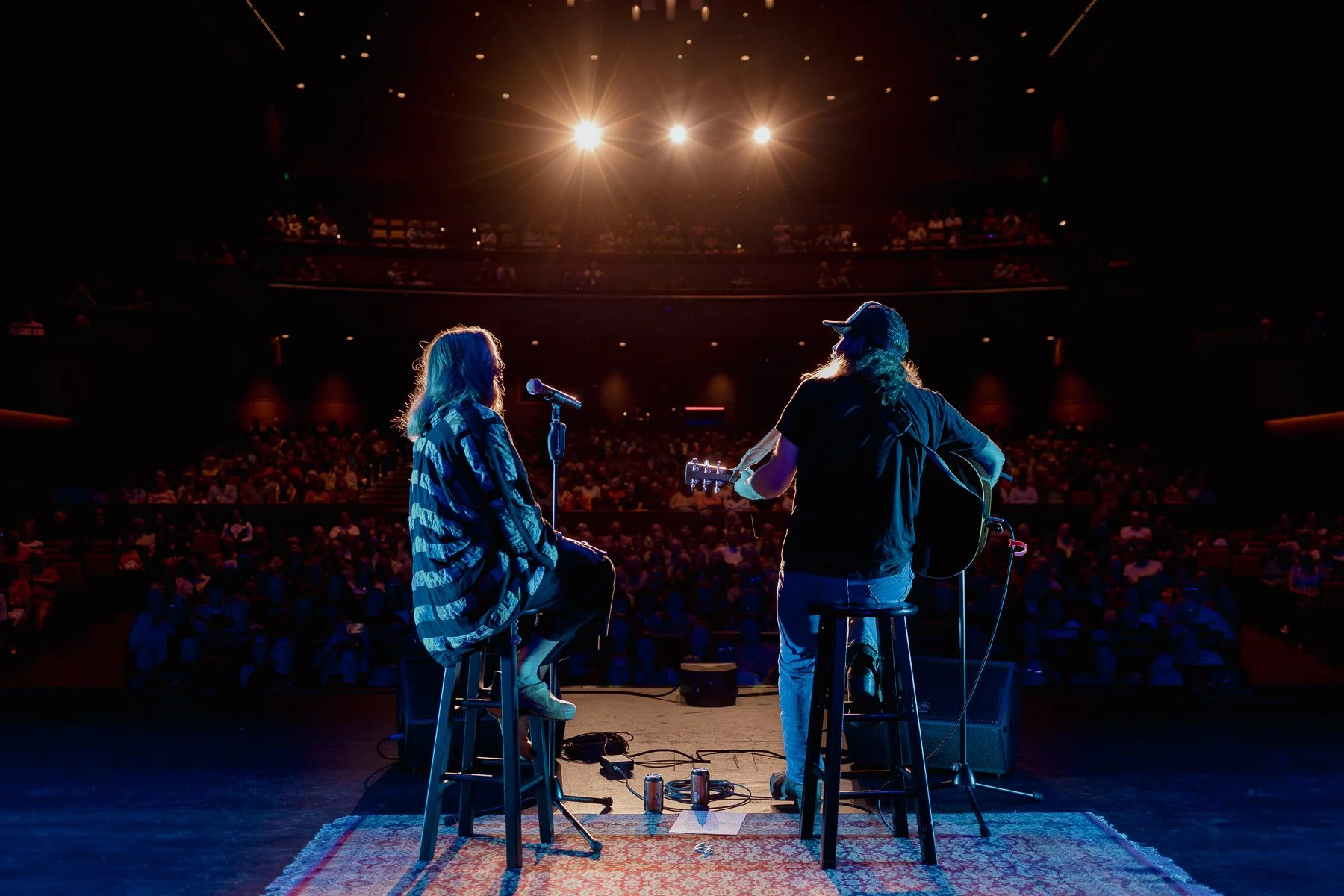 Two musicians on stage during a concert, one singing into a microphone and the other playing guitar with an audience in the background.