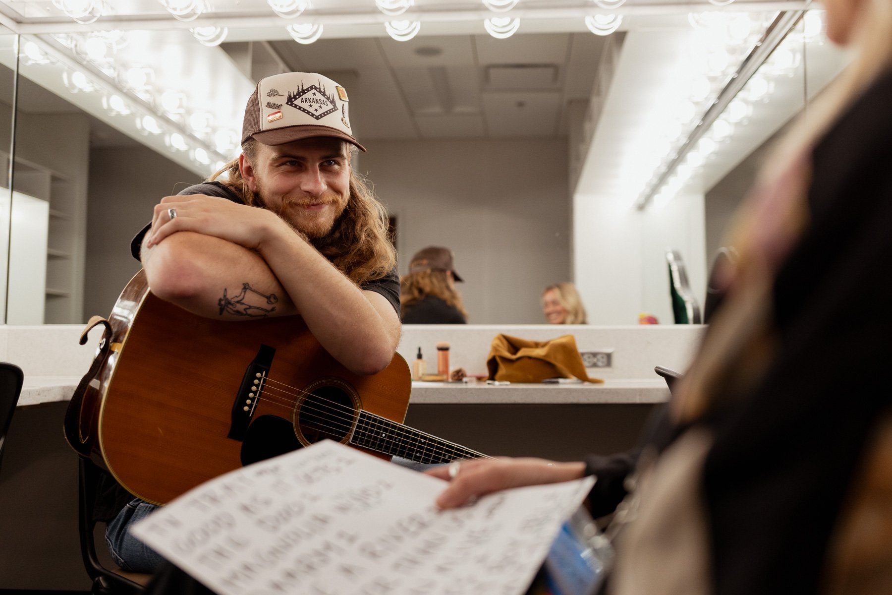 Man with Arkansas hat, with his hands folded over his guitar and a smile on his face