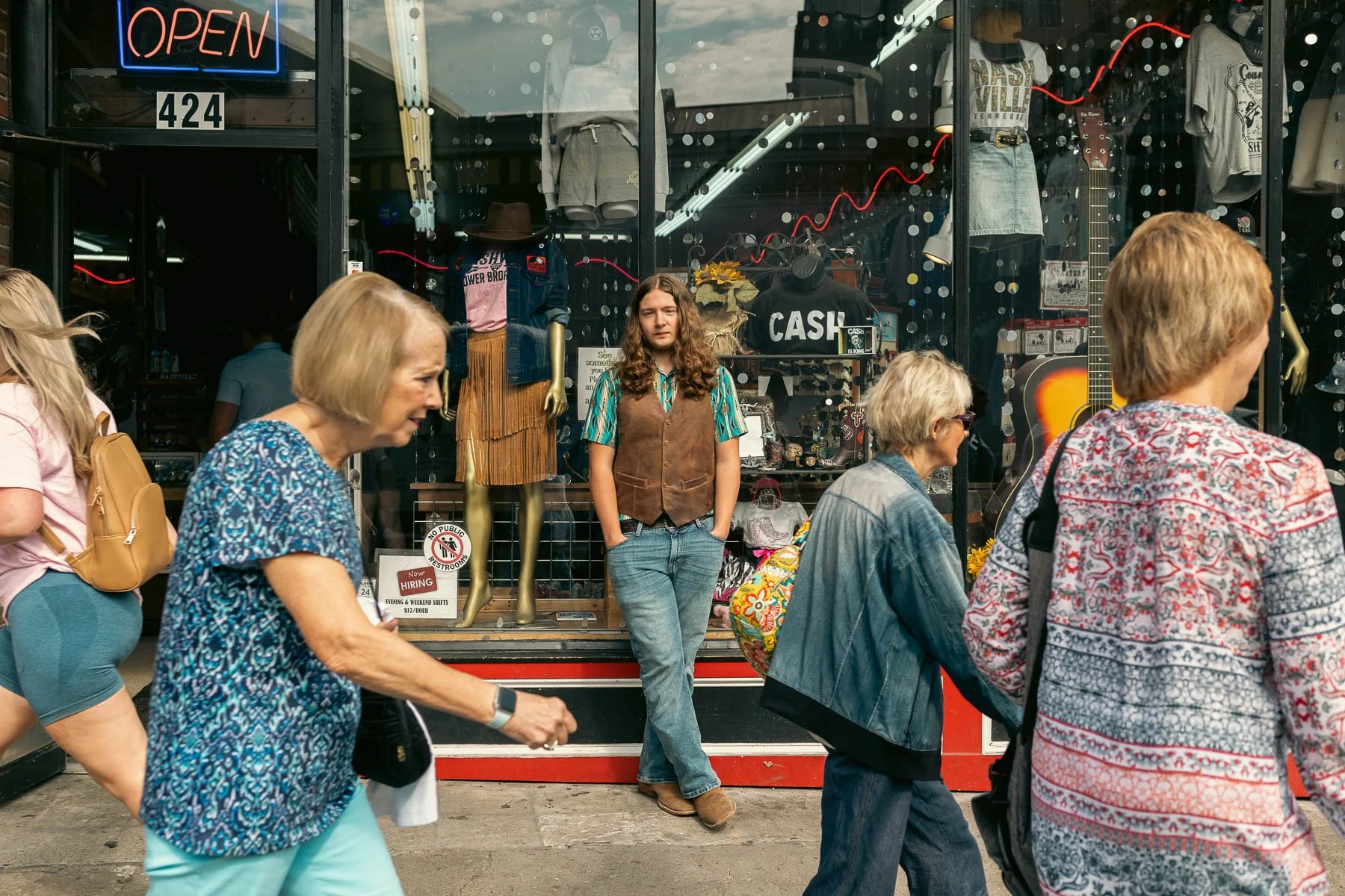 A young woman with brown hair and a floral shirt stands with her hands in her pockets in front of a shop window display, while pedestrians walk past on the street.
