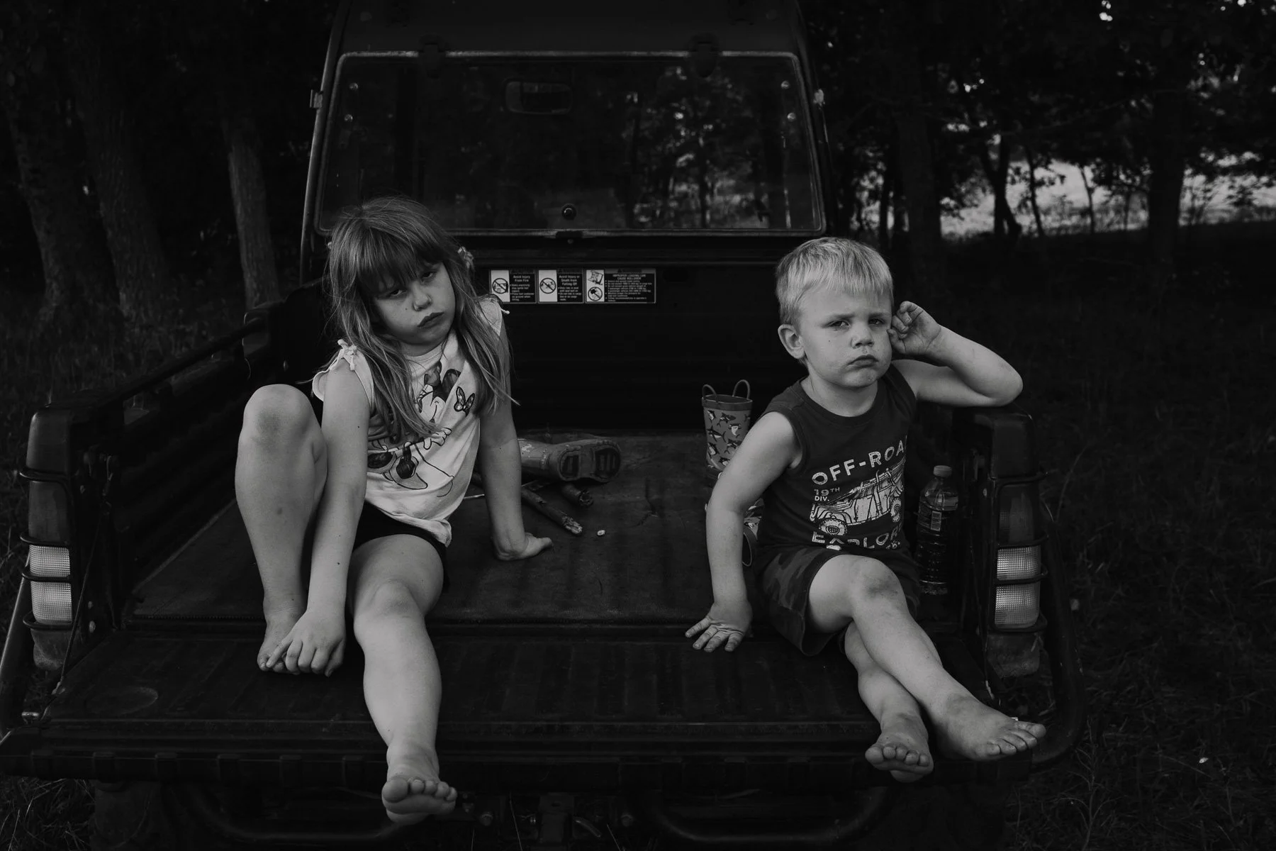 Two children sitting on the back of a pickup truck in a wooded area, looking at the camera with serious expressions.