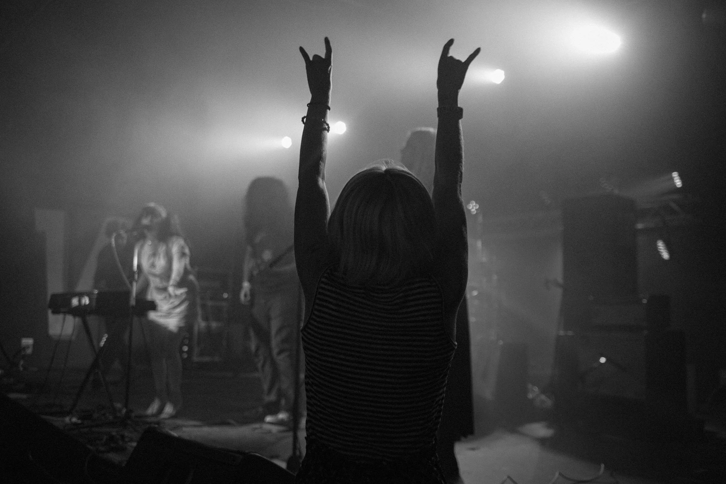 A person with long hair, wearing a striped sleeveless top, is raising their arms with hands making gesture in front of a stage with musicians, in a black and white photo.